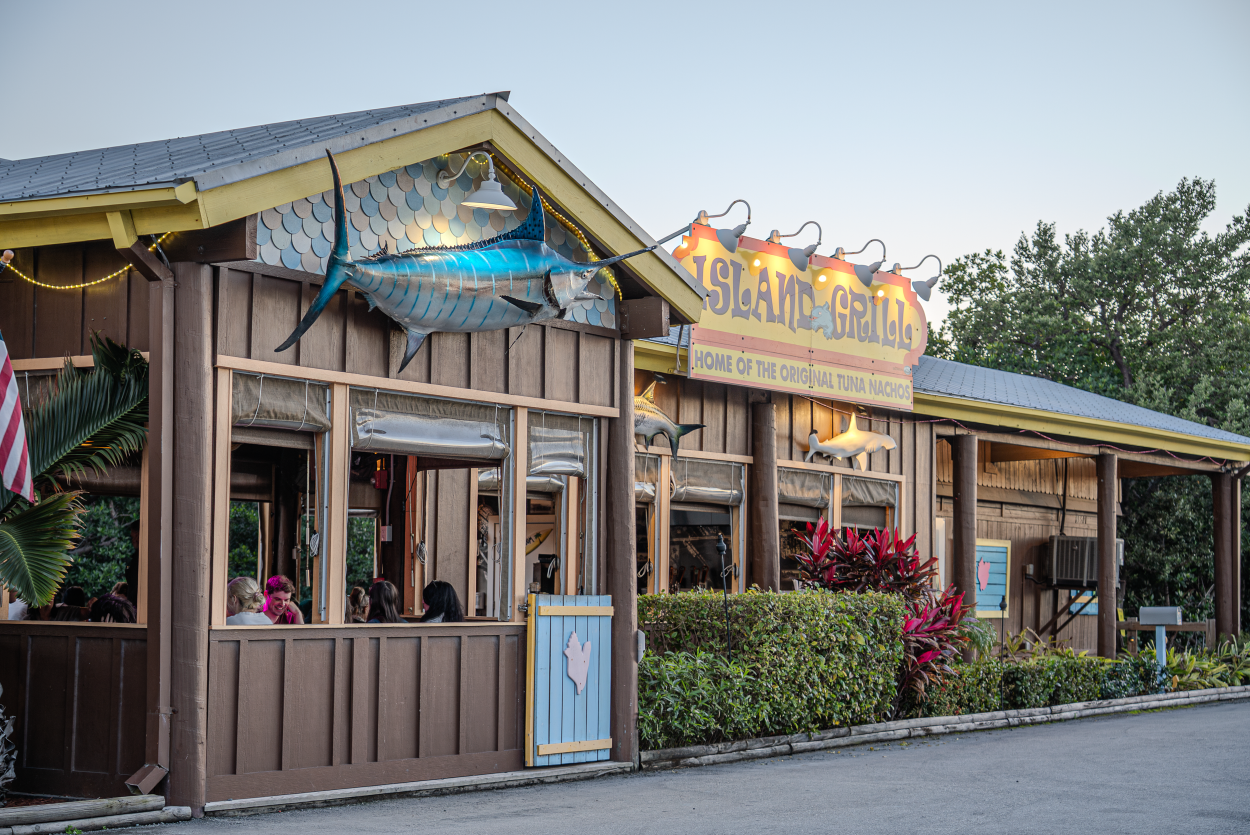 Outdoor restaurant named Island Grill with a large marlin fish mounted above the entrance, surrounded by trees and bushes, with people dining inside.
