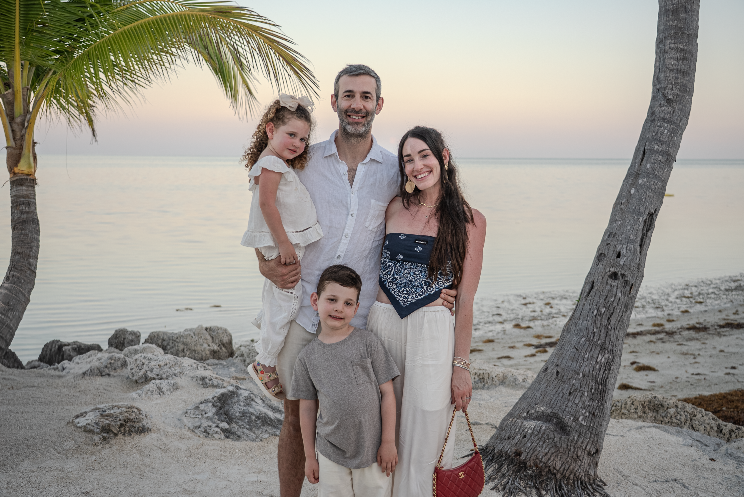 A smiling family of four standing on a beach with palm trees, rocks, and the ocean in the background during sunset.