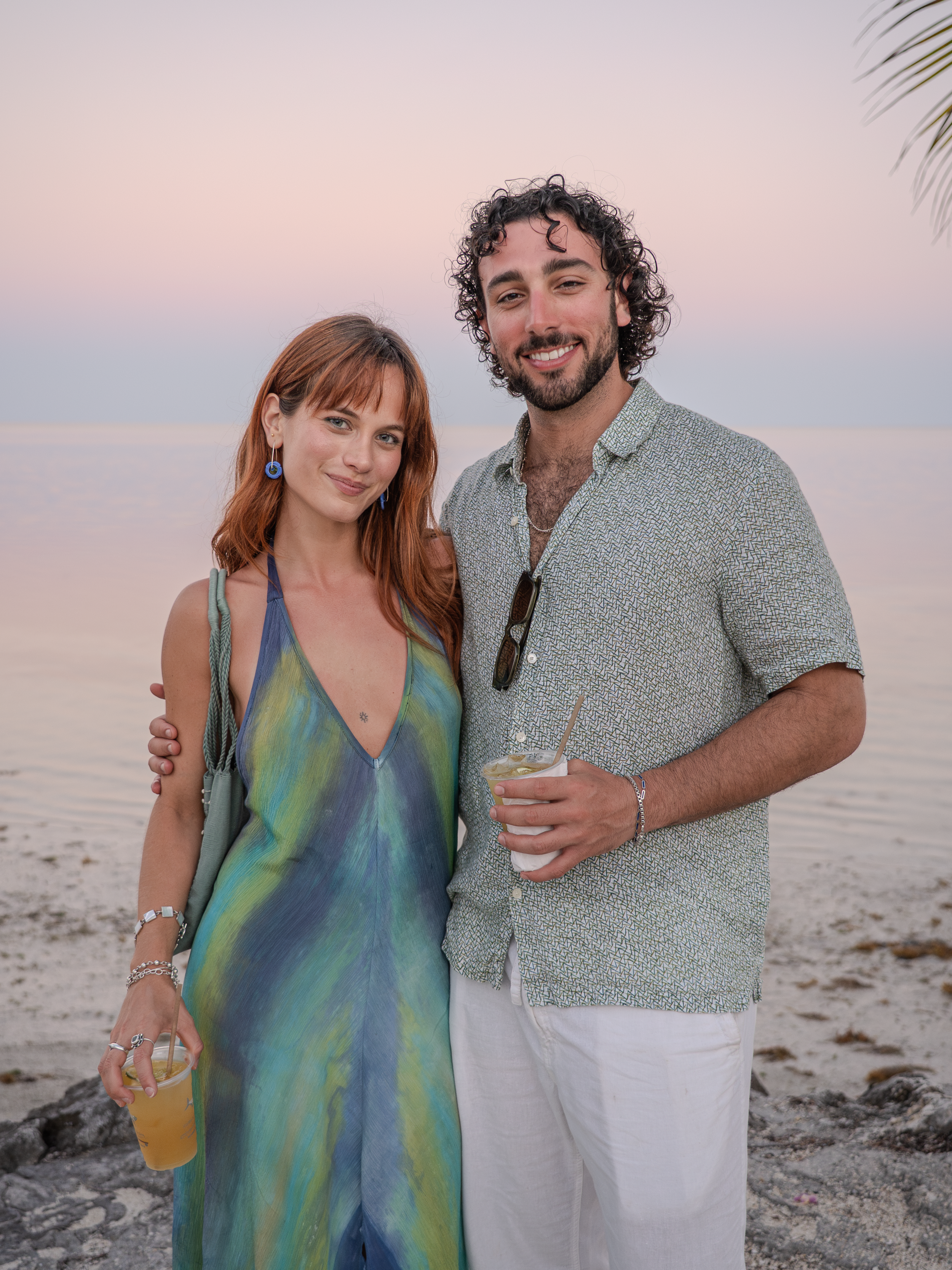 A young woman and man standing on a beach at sunset, smiling and holding drinks.