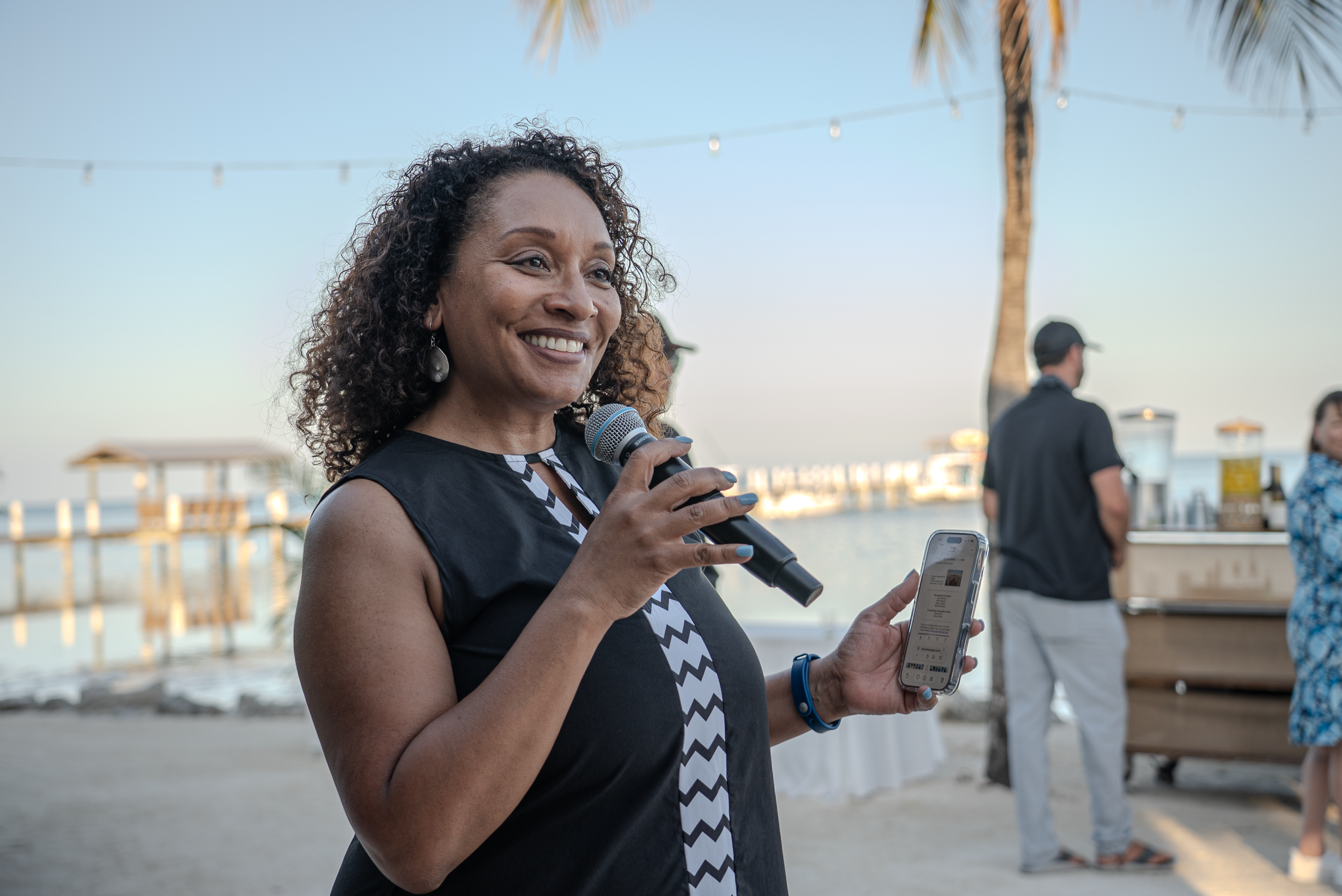A woman with curly hair and earrings holding a microphone in one hand and a smartphone in the other, standing outdoors near a beach with palm trees and string lights, during sunset.