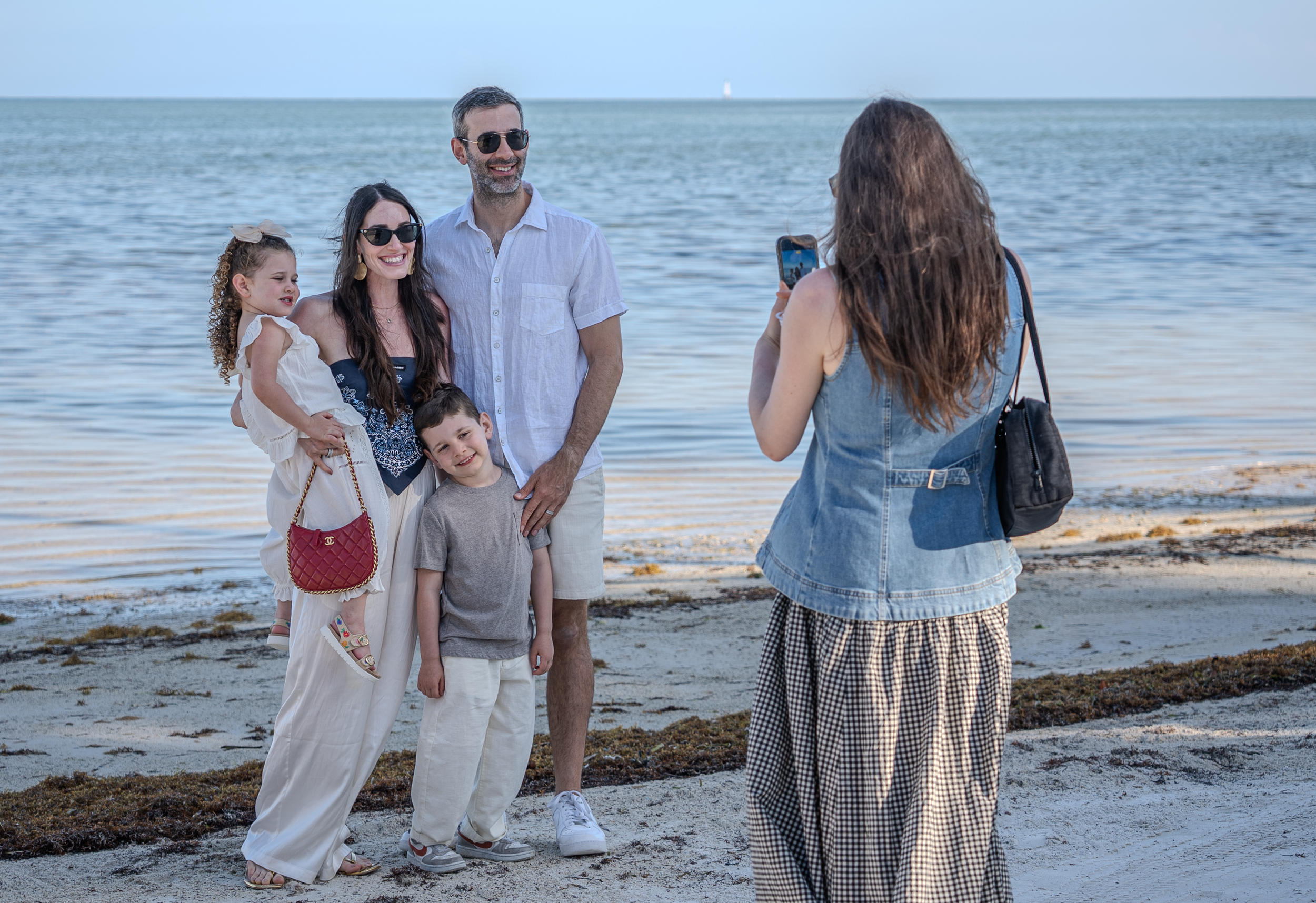 A family of five stands on a beach, being photographed by a woman with long brown hair, wearing a denim vest, holding a smartphone. The family includes two children, a woman, and a man, all smiling. The ocean and a lighthouse are visible in the background.