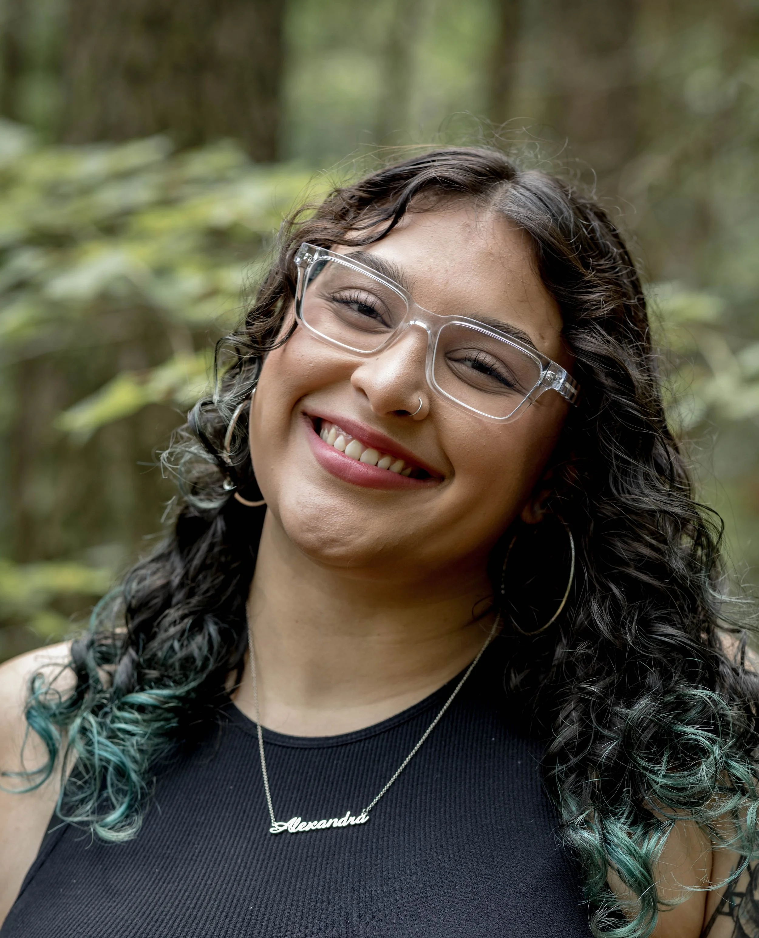Close-up of a smiling woman with curly hair, wearing clear glasses, a black top, and a silver necklace with a name pendant that says 'Alexandra', in an outdoor setting with blurred greenery in the background.