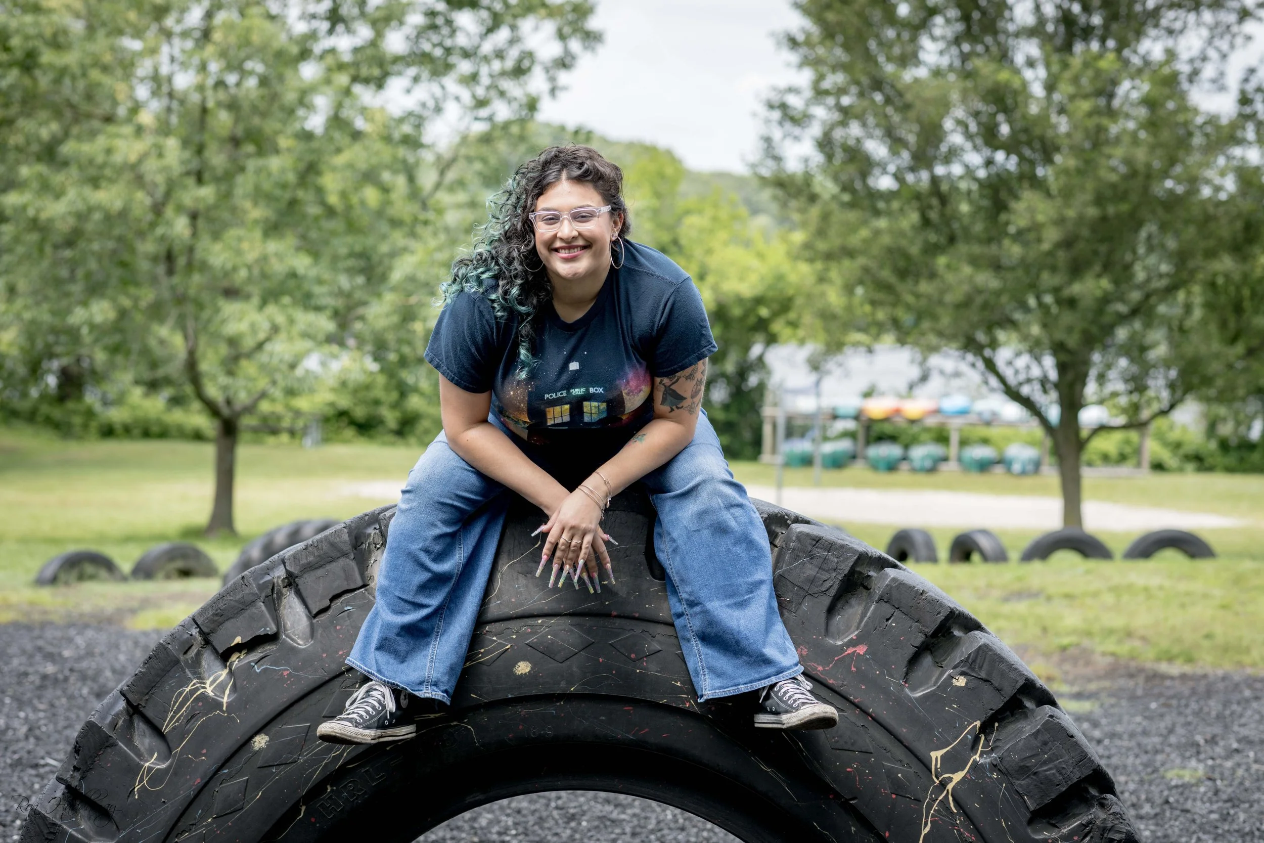 A young woman with curly hair, glasses, and tattoos smiling while sitting on an old tire in a park with trees in the background.