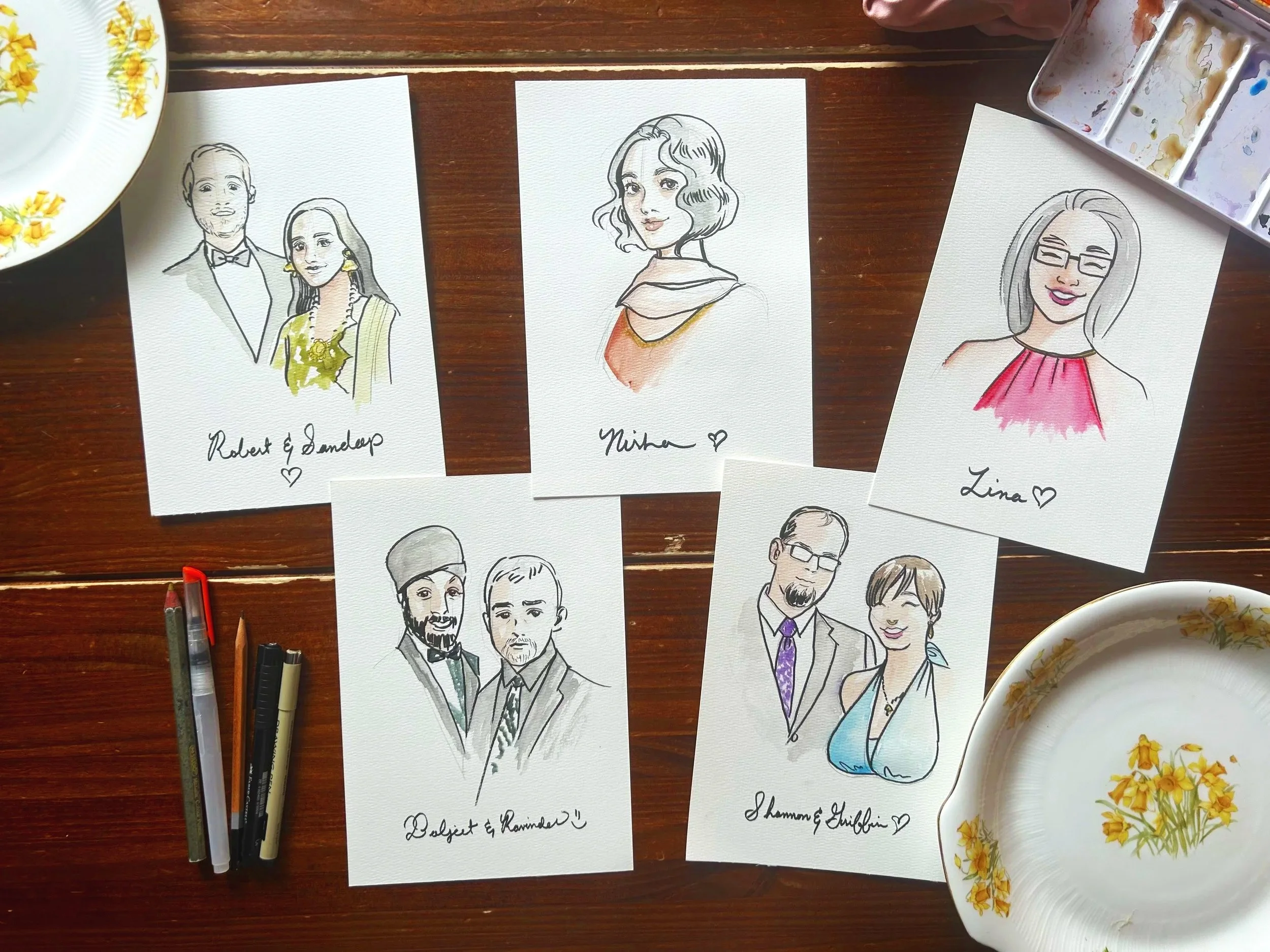 Set of hand-drawn caricature portraits of men and women on white paper, with some colored in and others in black and white, accompanied by handwritten names and hearts, on a wooden table with art supplies and plates nearby.