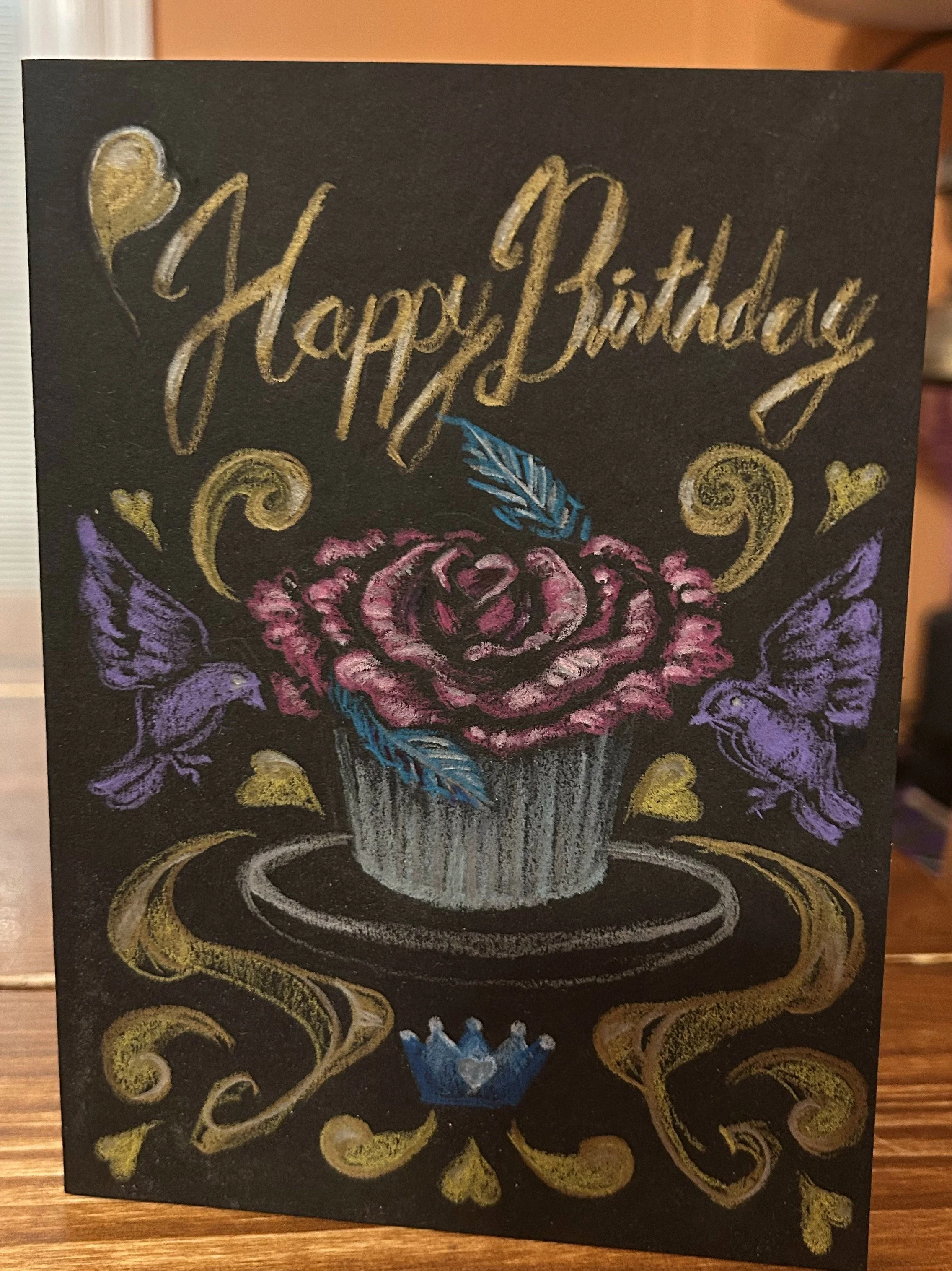 A chalk art birthday card with gold, pink, purple, blue, and white chalk colors on a black background. It features a floral design with the words "Happy Birthday!" written in gold cursive.