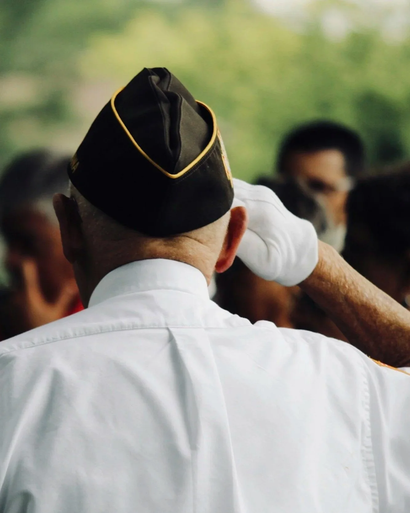 Close-up of a man in a white shirt saluting, with other people in the background, and a green blurred outdoor setting.