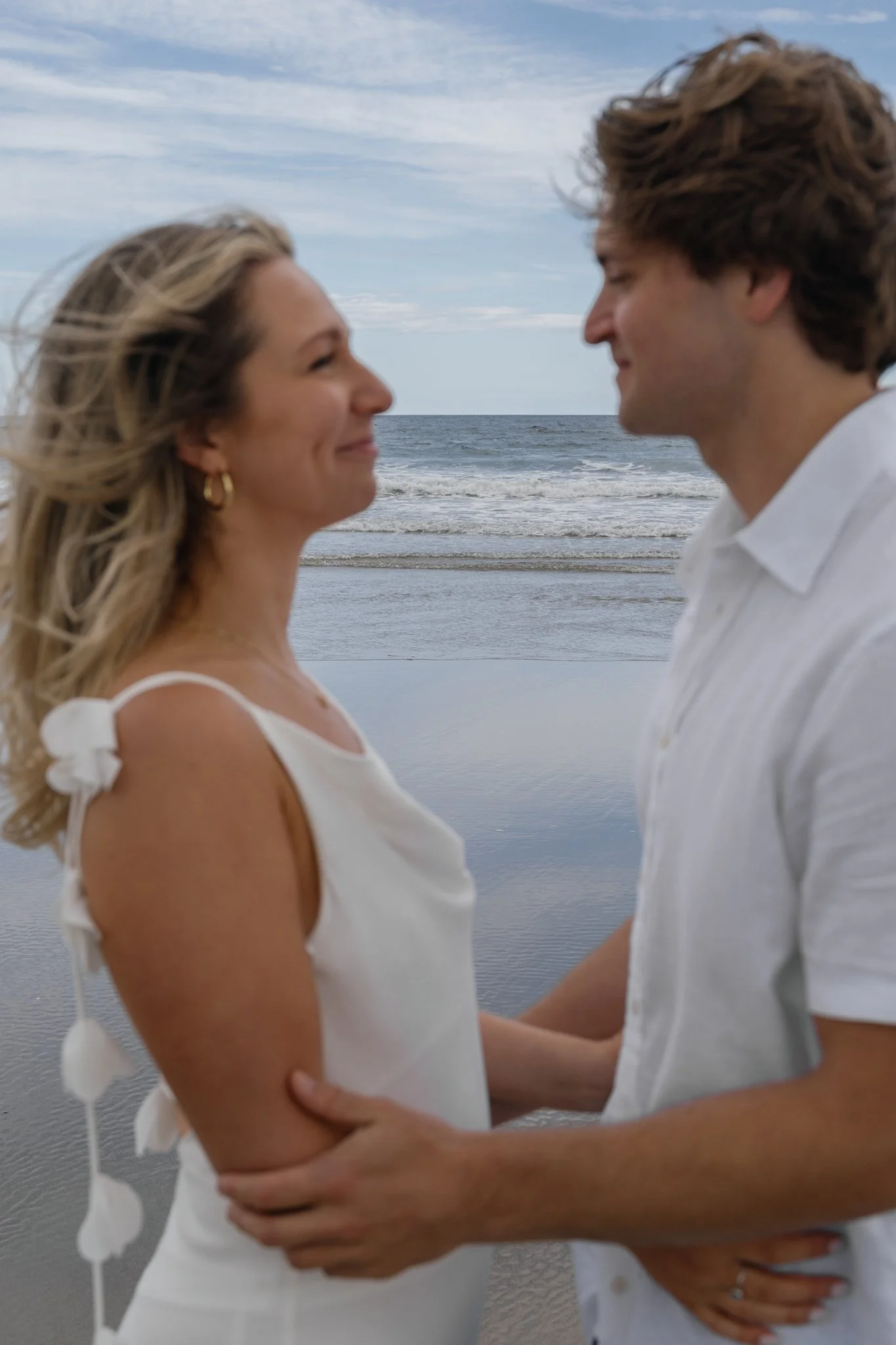 A couple holding hands and facing each other on a beach, with the ocean and a cloudy sky in the background.