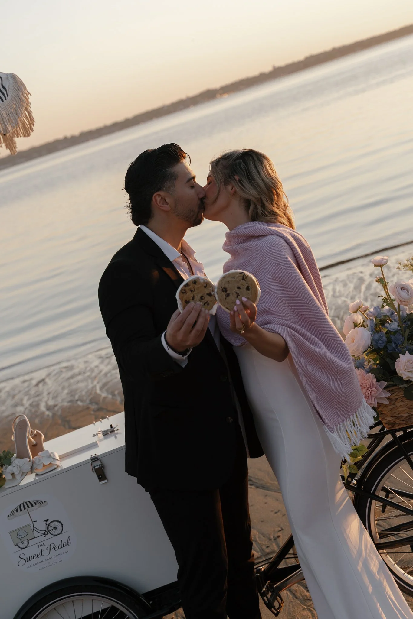 A couple kissing on the beach during sunset, holding cookies, with an ice cream cart nearby.