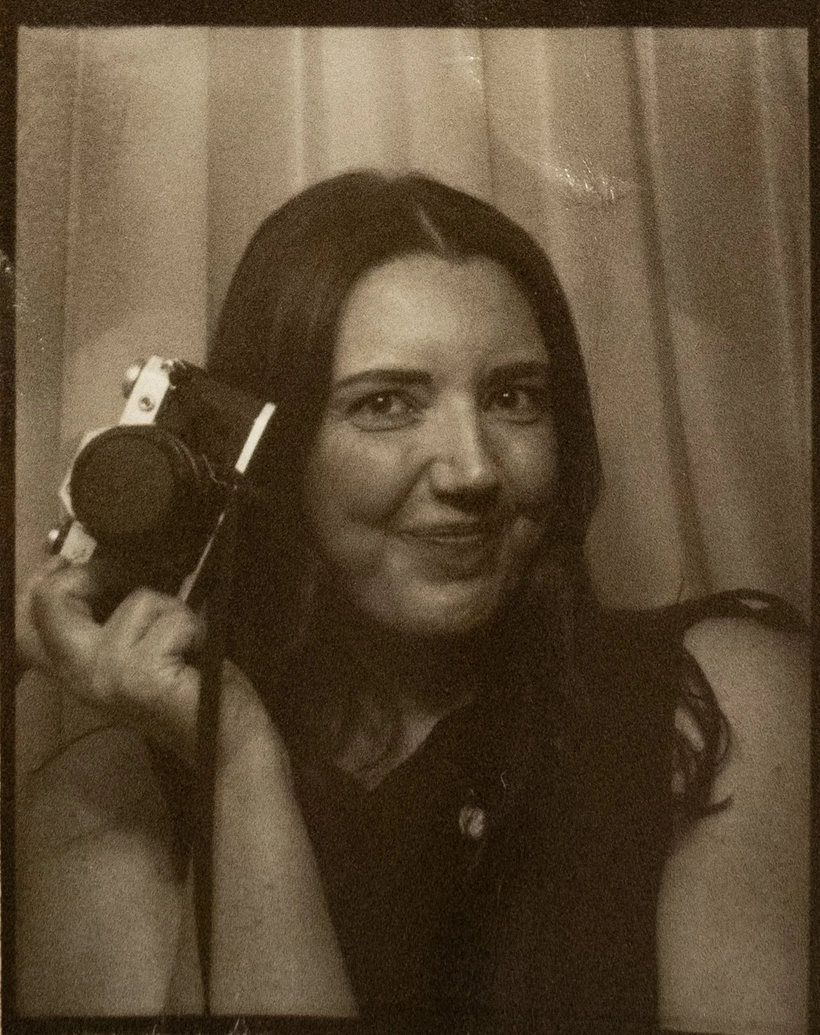 A woman with light skin and dark hair, using headphones and holding a microphone in front of her mouth, smiling and looking at the camera.