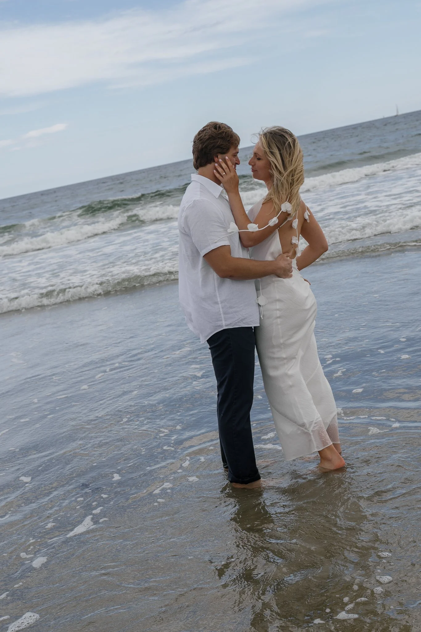 A couple standing in shallow ocean water, looking into each other's eyes, with waves and a cloudy sky in the background.