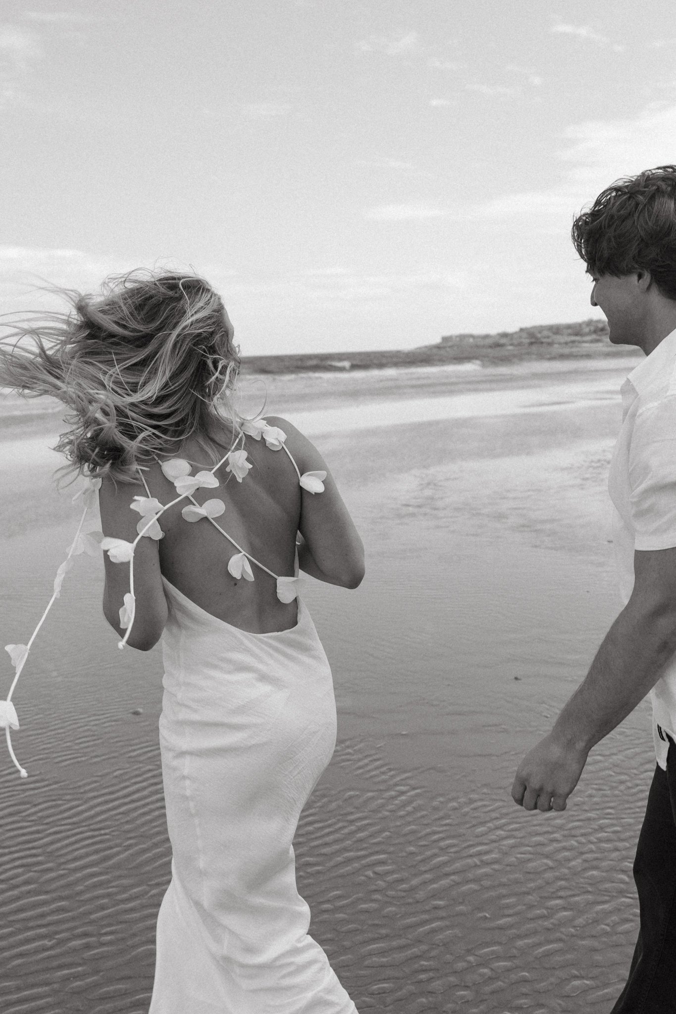 A couple holding hands on the beach, with the woman wearing a dress with leaves, and the man in a white shirt, holding hands, with ocean and distant land in the background.
