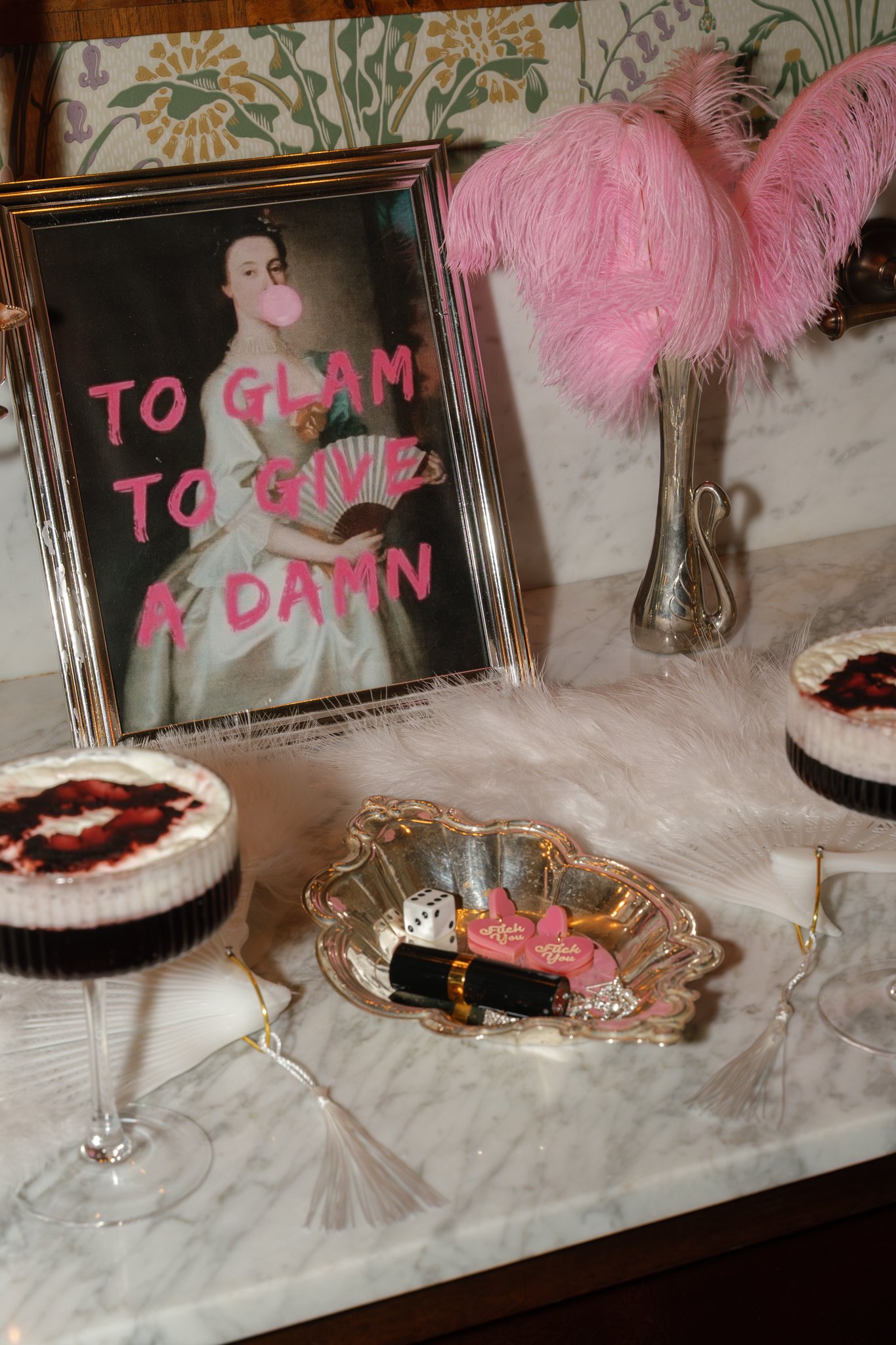 A decorated vanity table with pink feathered accessories, a framed vintage portrait with pink text, and with small containers of desserts, lipsticks, dice, and jewelry on a marble surface.