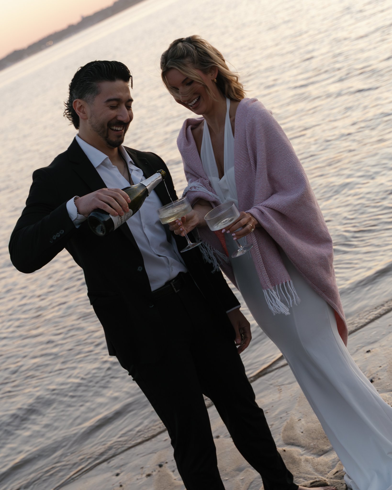 A man and woman stand close together on a beach at sunset, smiling and raising glasses for a toast. The man, in a black suit, pours champagne into a glass held by the woman, who wears a light pink shawl and white outfit. The background features calm 