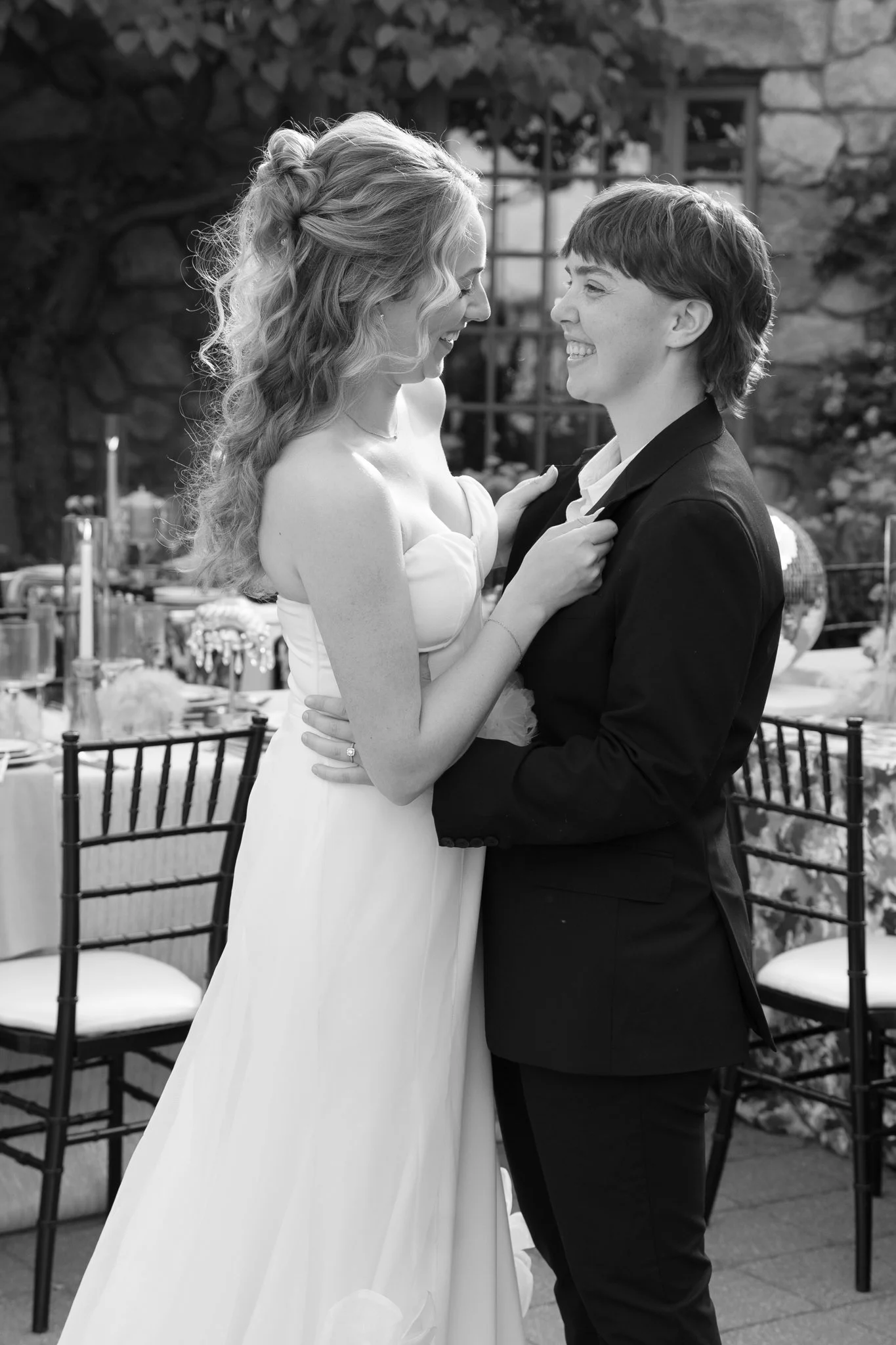 A black-and-white photo of a happy couple at their wedding, standing close and smiling at each other. The bride is in a strapless wedding dress with long, wavy hair, and the groom is in a tuxedo with a white shirt and dark jacket, holding her hand.