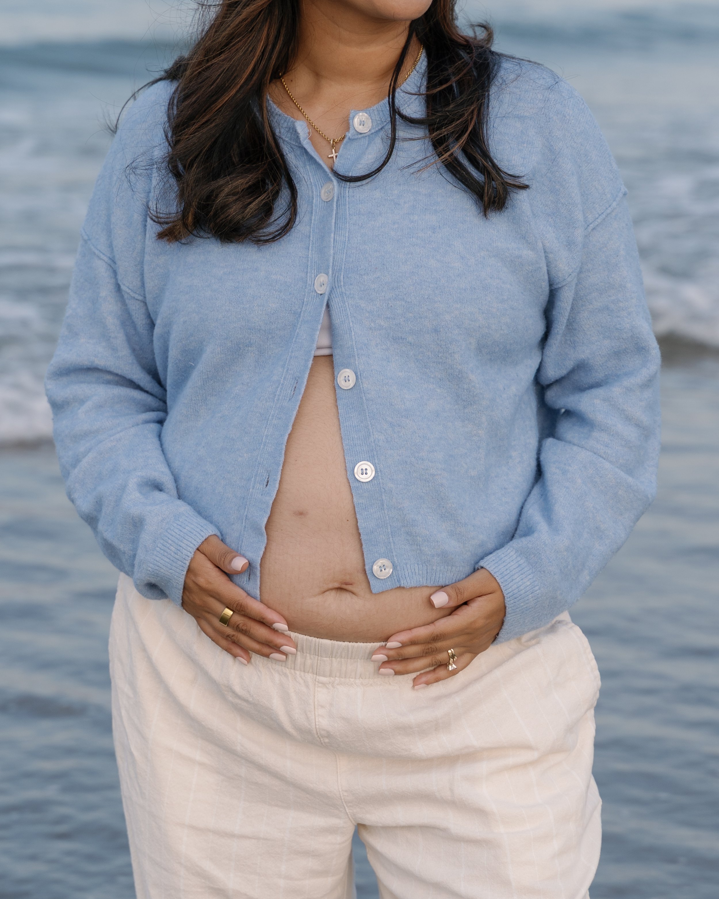 A woman wearing a light blue cardigan, standing near the ocean with her hands resting on her abdomen, wearing rings and a necklace with a cross pendant.