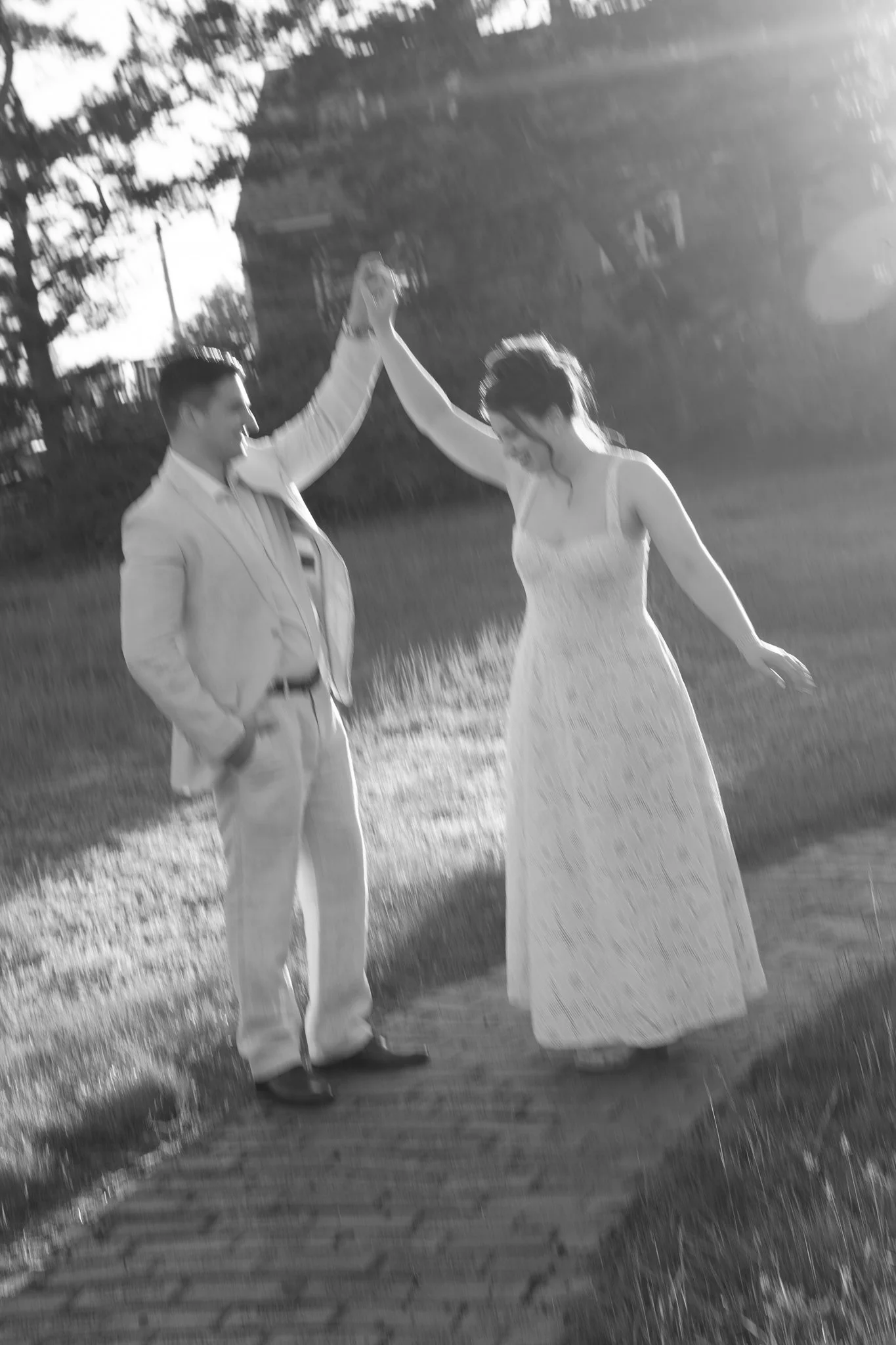 A black and white photo of a couple dancing outdoors by a body of water, holding hands and smiling, with trees in the background.