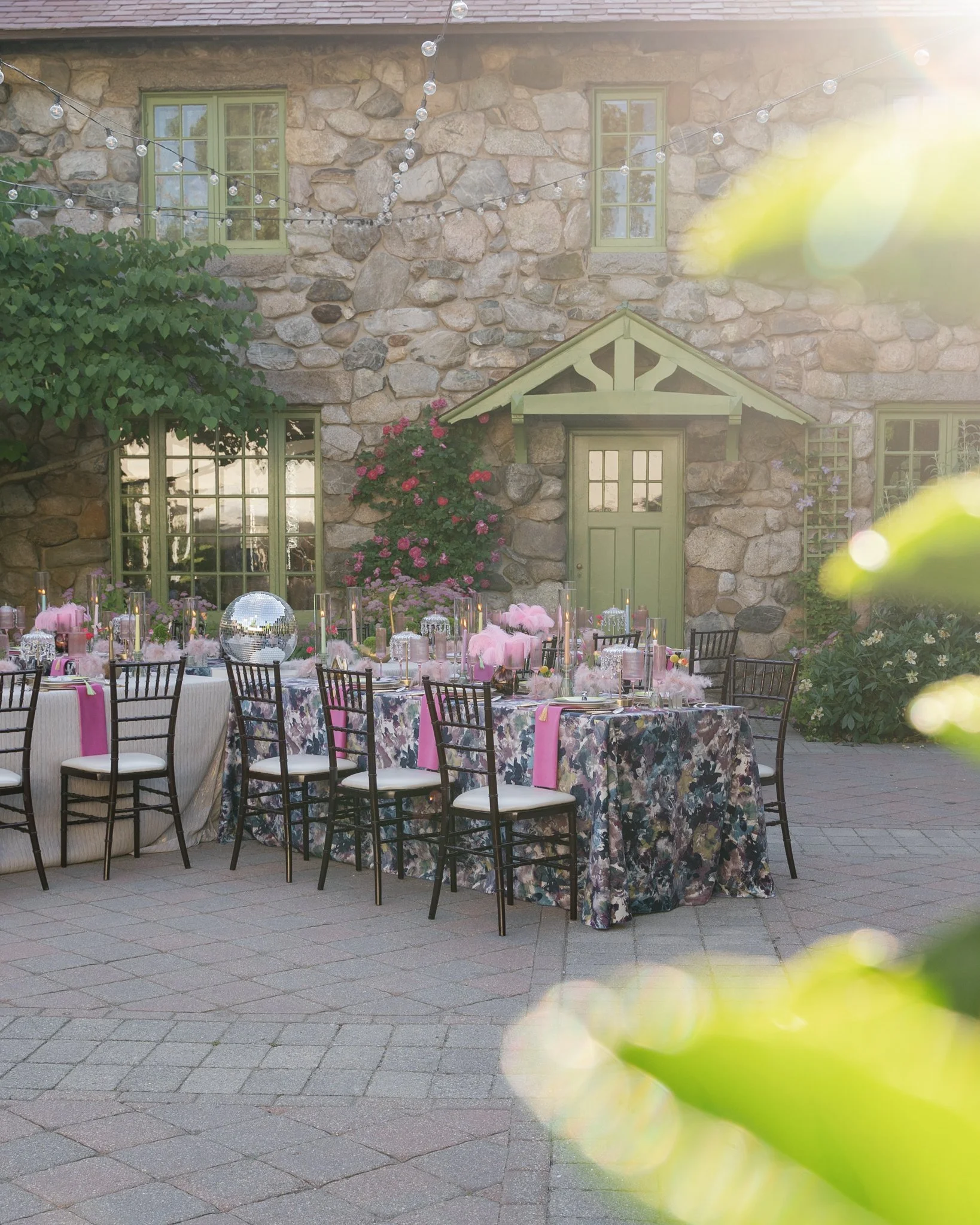 Outdoor dining setup with decorated tables, chairs, and a stone building in the background, with string lights hanging overhead.
