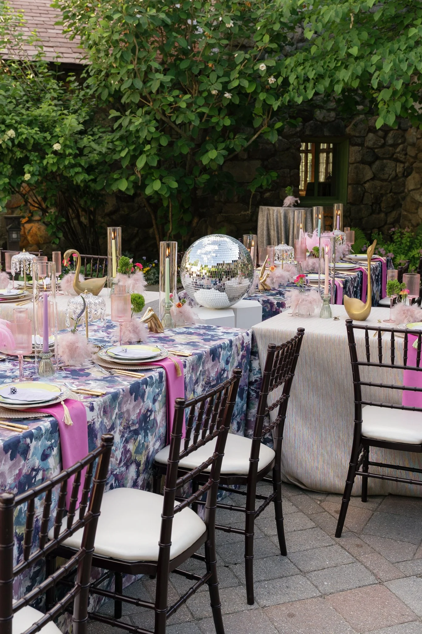 Outdoor party setup with tables decorated with pink and purple tablecloths, pink candles, gold swan sculptures, disco ball, pink feathers, and chairs around the tables. Green plants and stone wall in the background.