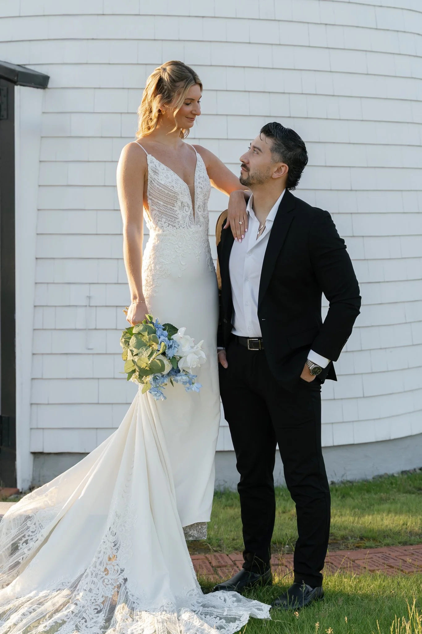 A bride and groom are standing outdoors in front of a white wall. The bride is wearing a white wedding dress with lace details and holding a bouquet of flowers. The groom is wearing a black suit with a white shirt and has his hand on his hip while looking at the bride. The bride has her hand resting on the groom's shoulder and is looking down at him with a gentle smile.
