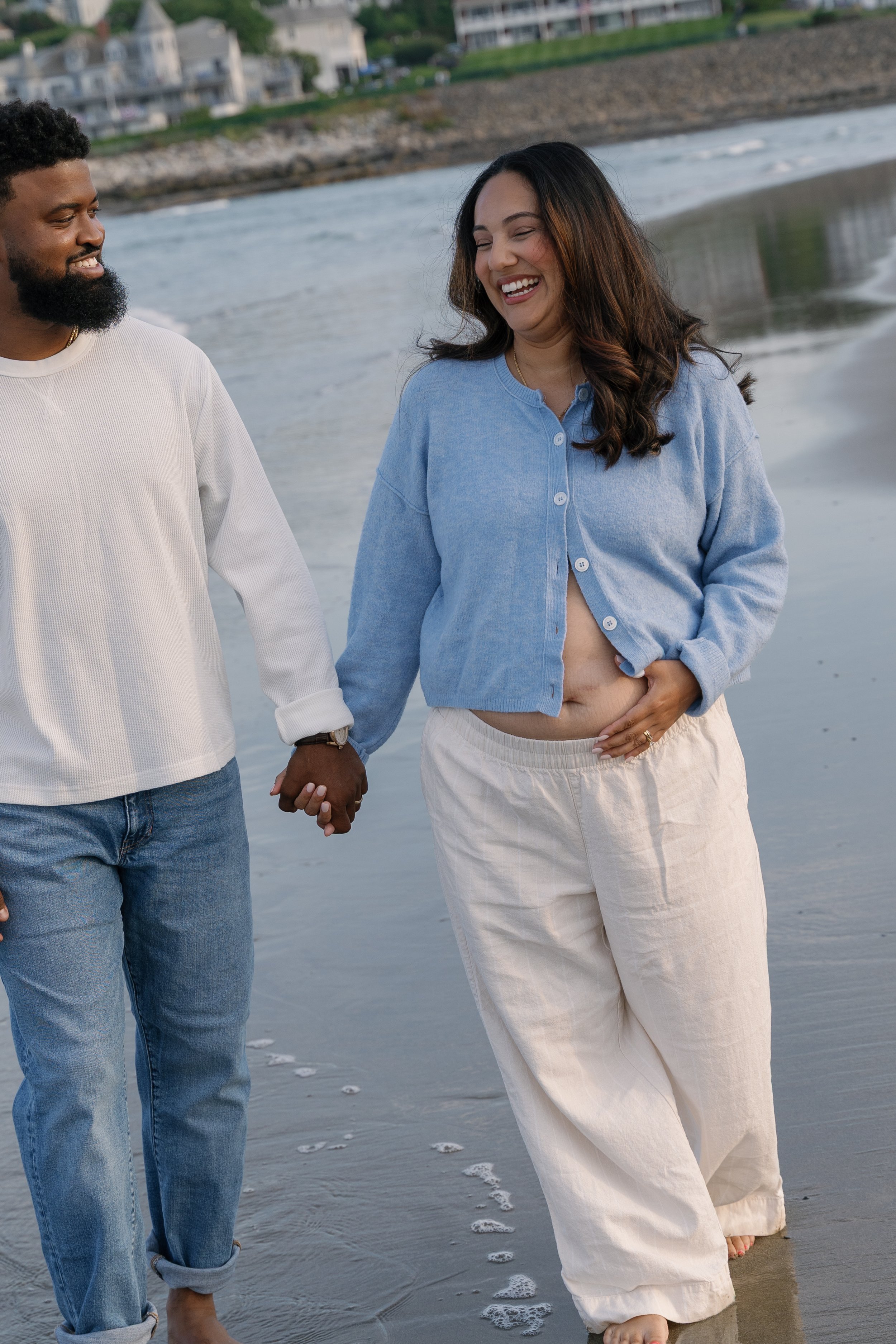 A smiling couple holding hands and walking along the beach, with the woman slightly lifting her dress and the man wearing a white long sleeve shirt and jeans, small waves touching their feet, houses in the background.