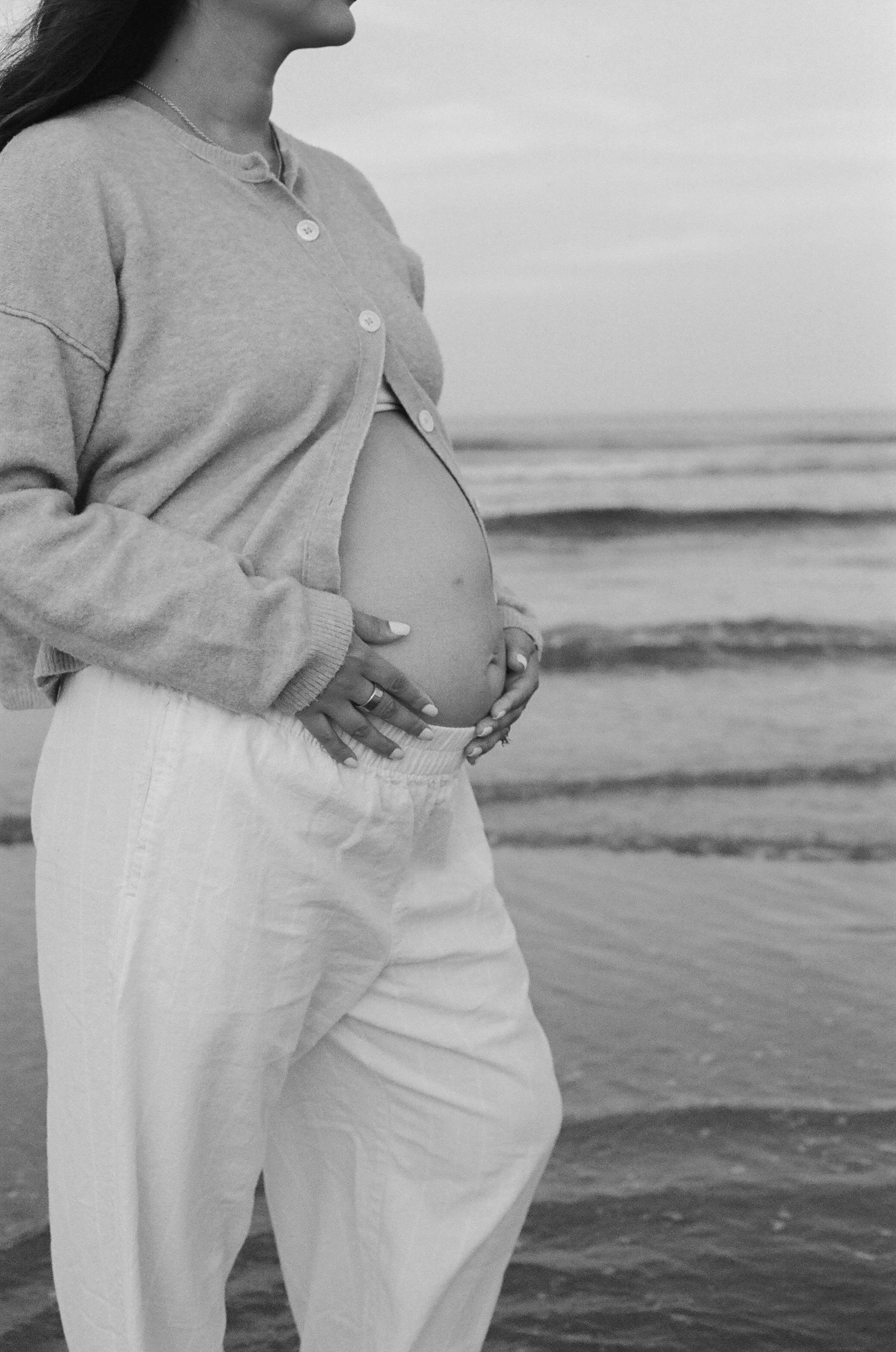 Close-up of a pregnant woman holding her belly while standing near the ocean, with waves in the background.