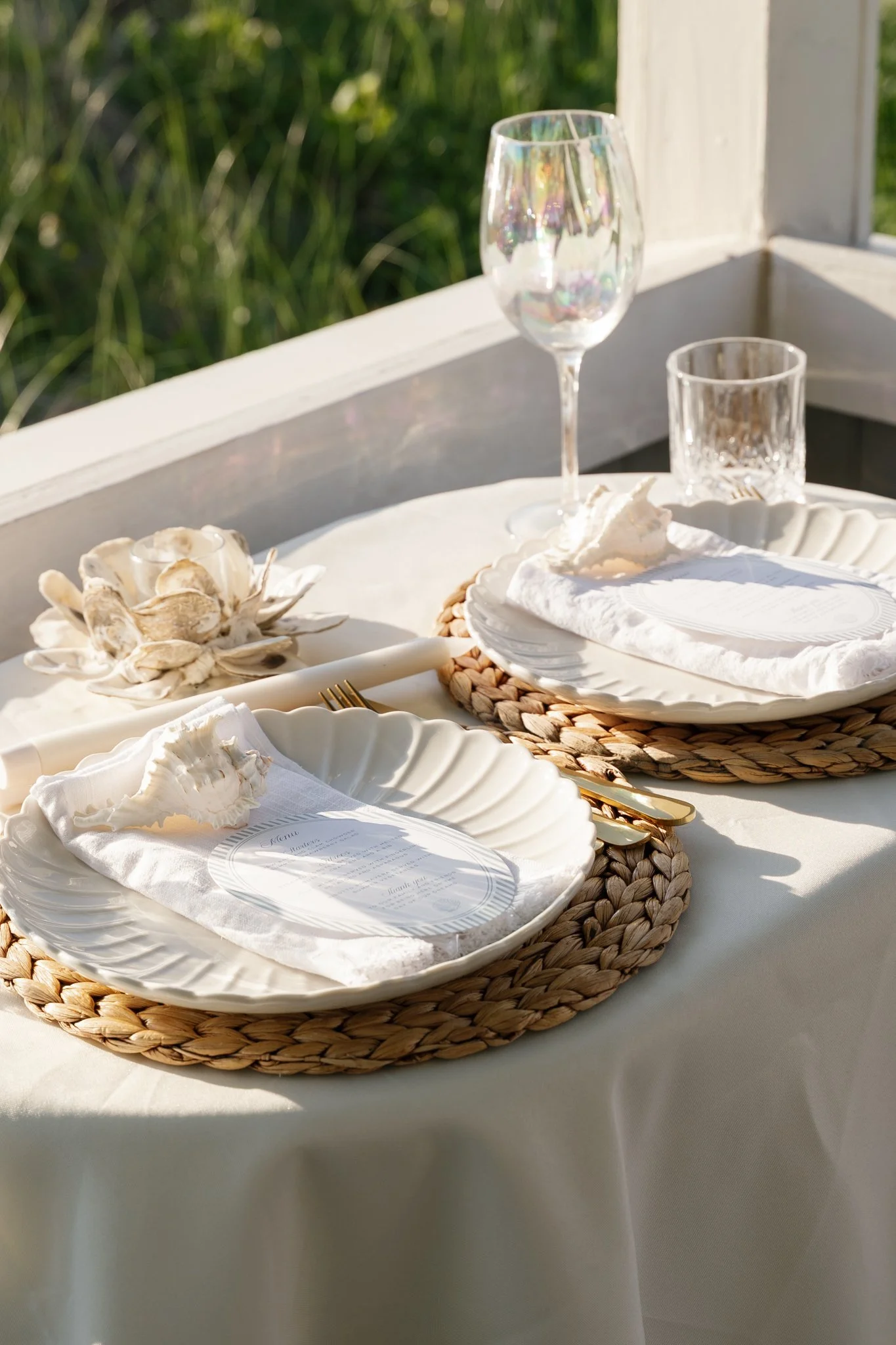 A table with white plates, cloth napkins, and gold utensils, set outdoors with seashell decorations, glassware, and a placemat on a white tablecloth.