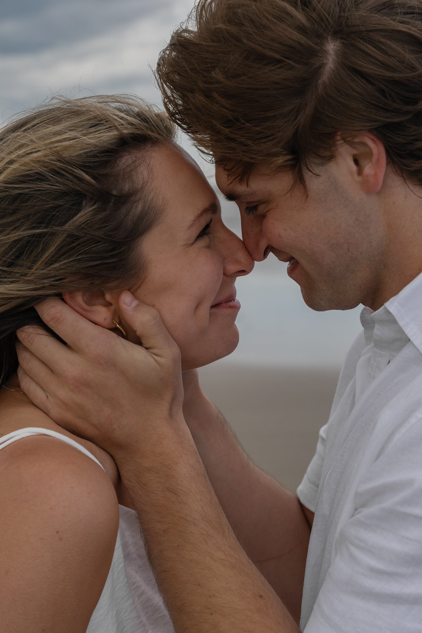 A man and woman are about to kiss each other with their faces close, smiling, outdoors under a cloudy sky.