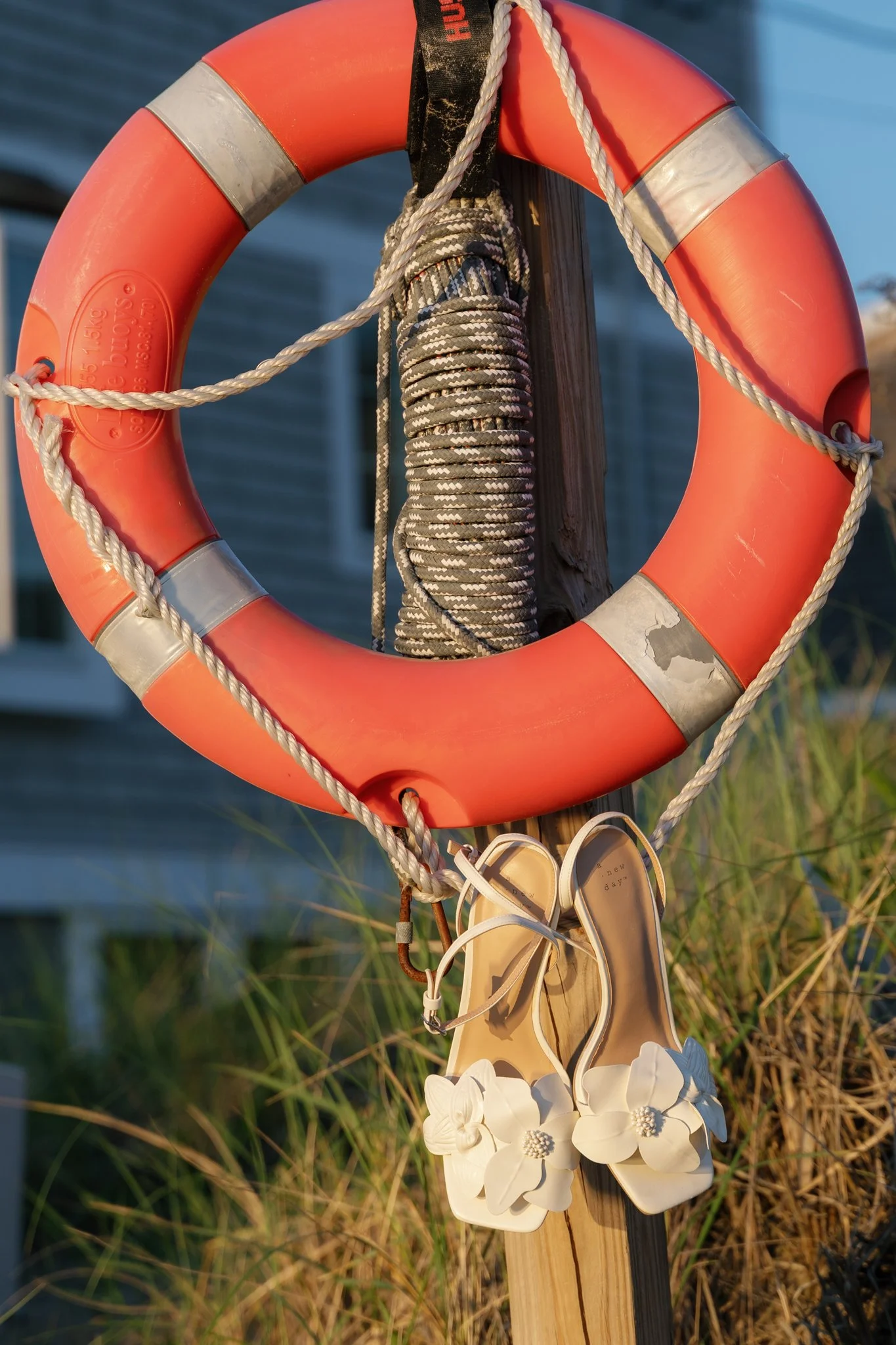 A life preserver ring attached to a wooden post with a black rope and a pair of white sneakers hanging at the bottom. There are some white flowers hanging on the post as well. The background shows water and green grass.