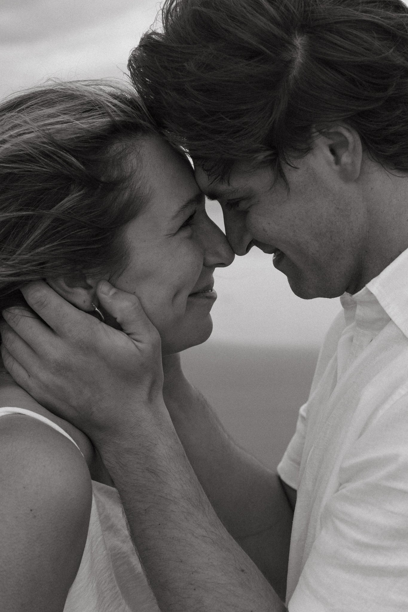 A close-up black and white photo of a couple touching foreheads with eyes closed, intimate and tender moment, both smiling gently, with one person's hand cradling the other's face.