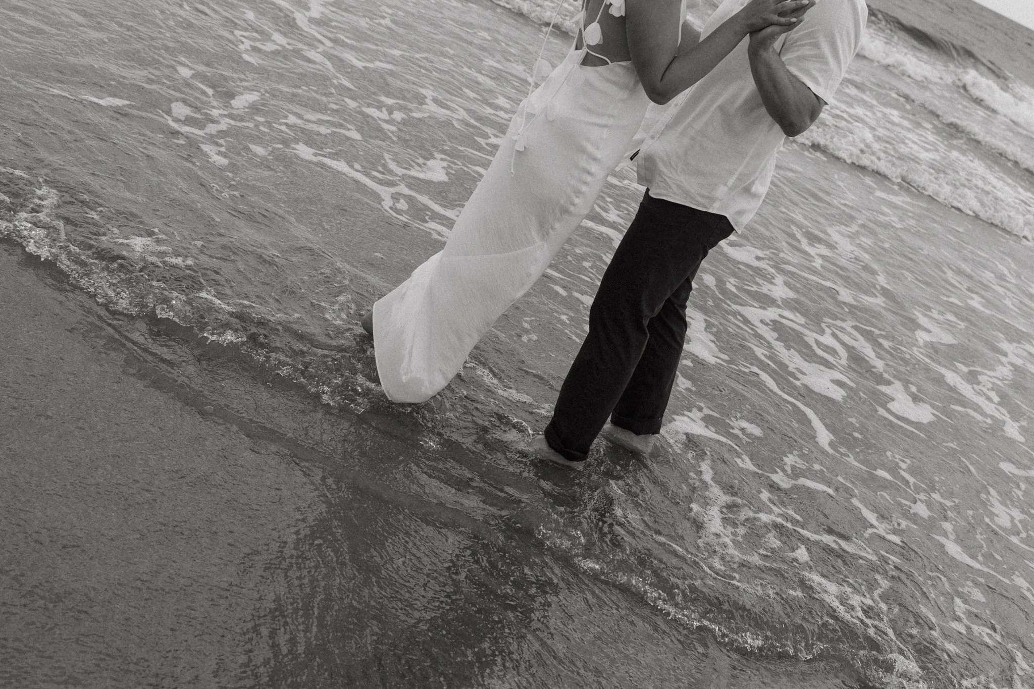 A couple holding hands and sharing a kiss at the beach with the ocean waves in the background.