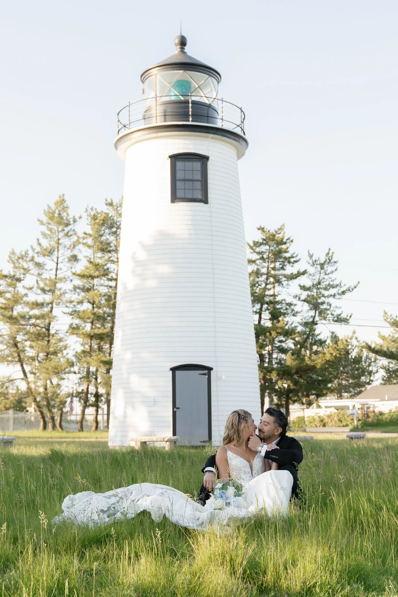A couple in wedding attire sitting on grass in front of a white lighthouse, sharing a kiss surrounded by trees.