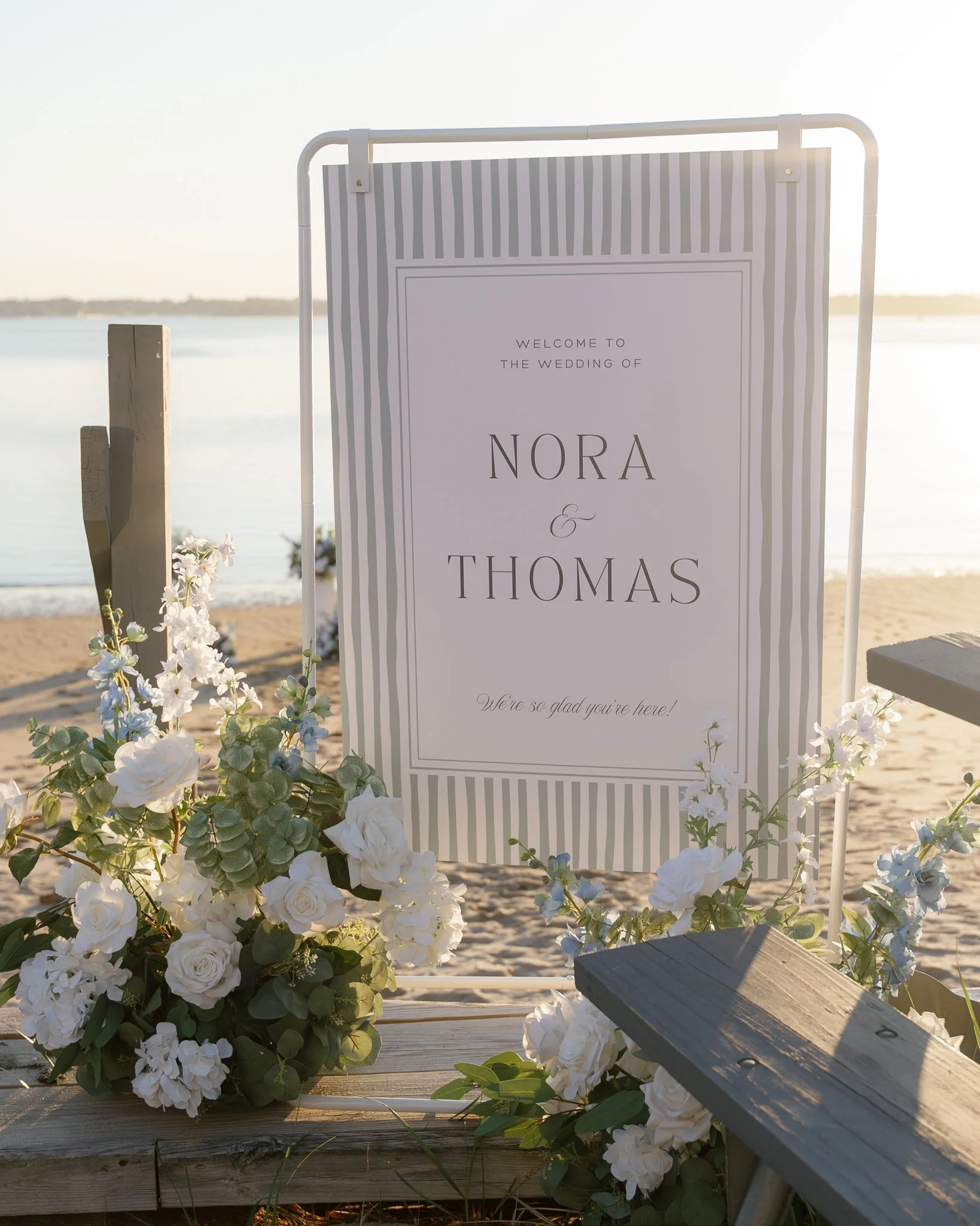 A wedding sign reading "Welcome to the wedding of Nora & Thomas" surrounded by white flowers and greenery, with a beach and water in the background.