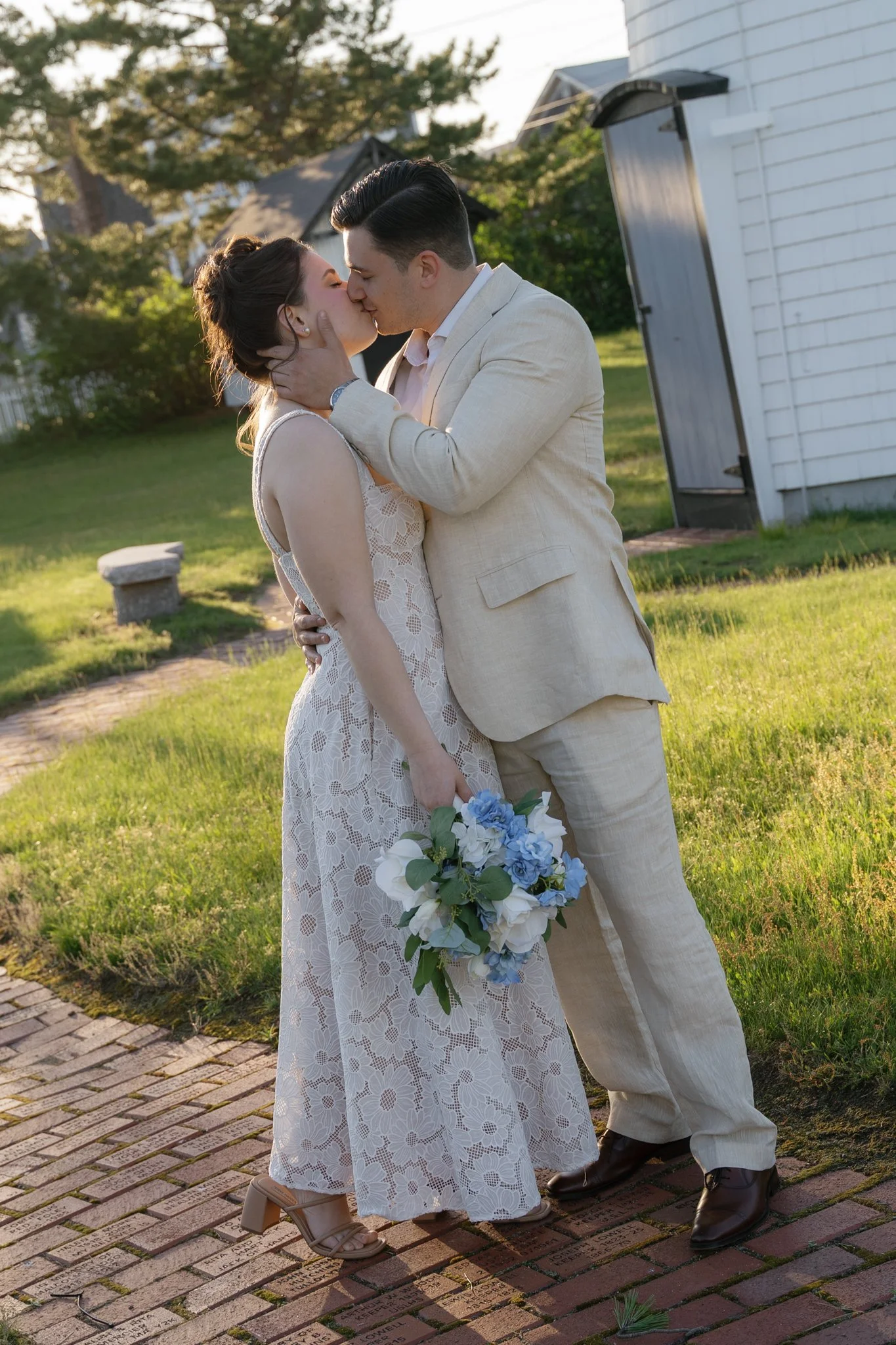 A couple in wedding attire sharing a kiss outdoors, with the woman holding a bouquet of white and blue flowers, on a brick path with engraved stones.