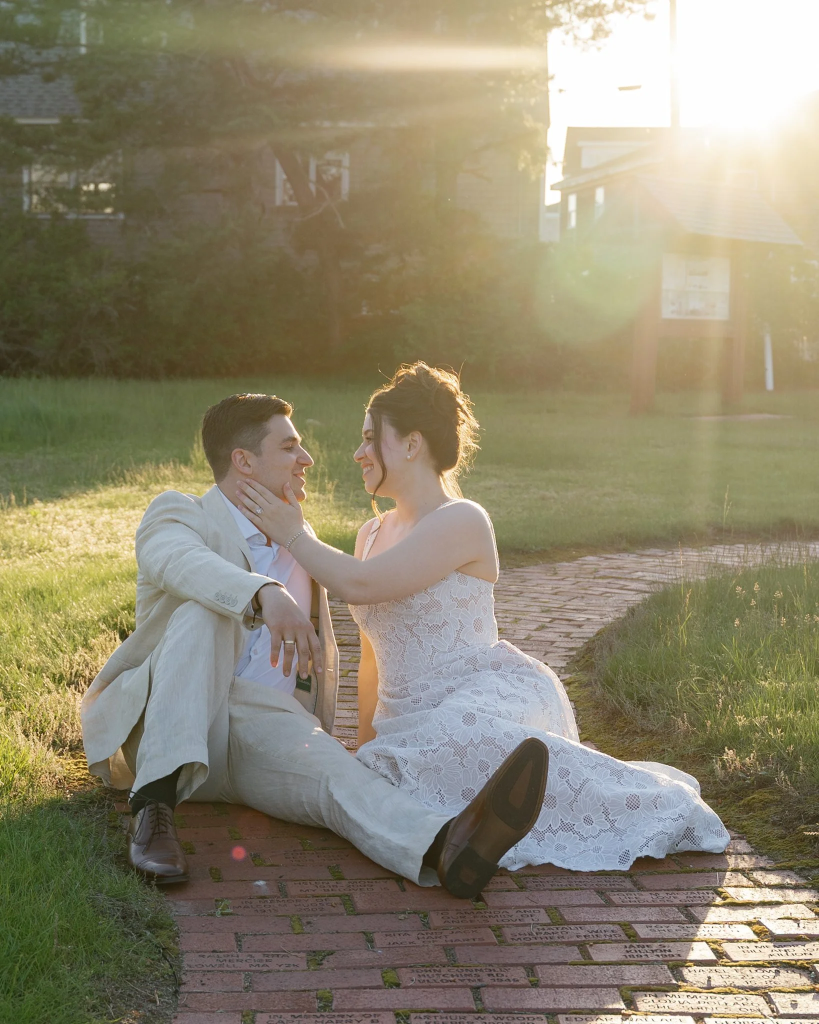 A couple dressed in wedding attire sitting on a brick pathway, gazing at each other with the sun setting behind them, creating a warm glow.