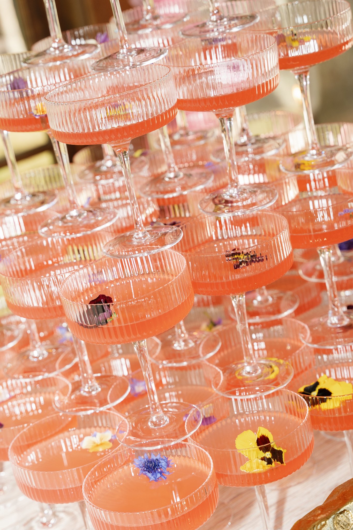 Multiple pink cocktails in tall, ribbed glasses with slices of yellow and purple edible flowers floating on the surface, arranged in a pyramid display.