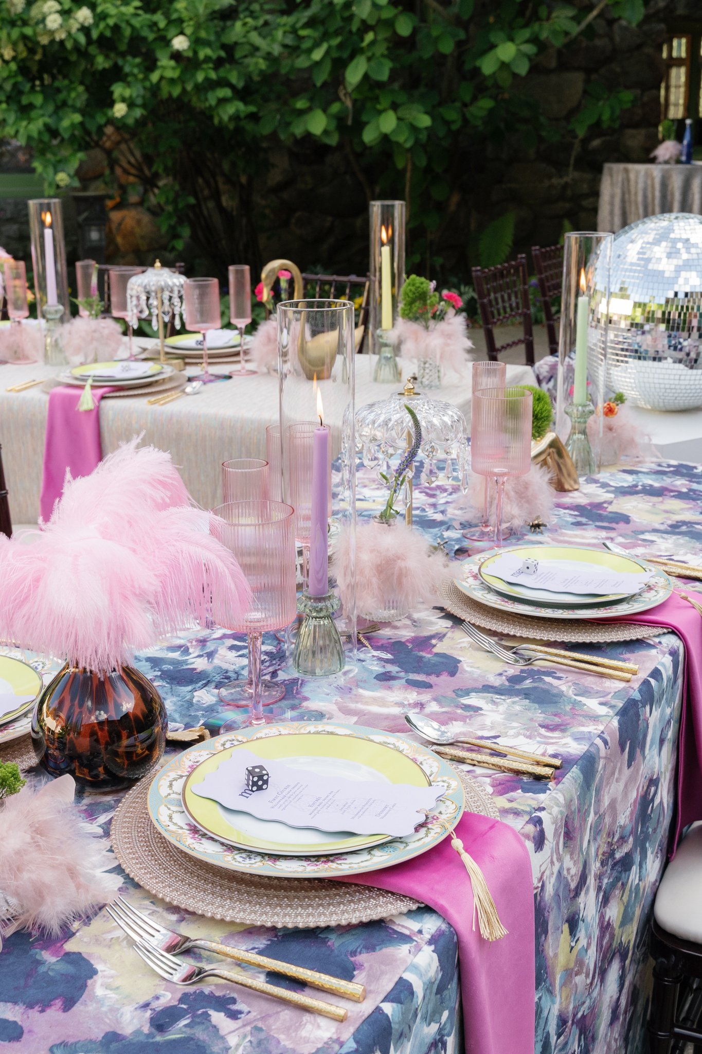 A beautifully decorated outdoor table set with pink feather accents, glassware, candles, floral arrangements, and a colorful tablecloth for a festive occasion.