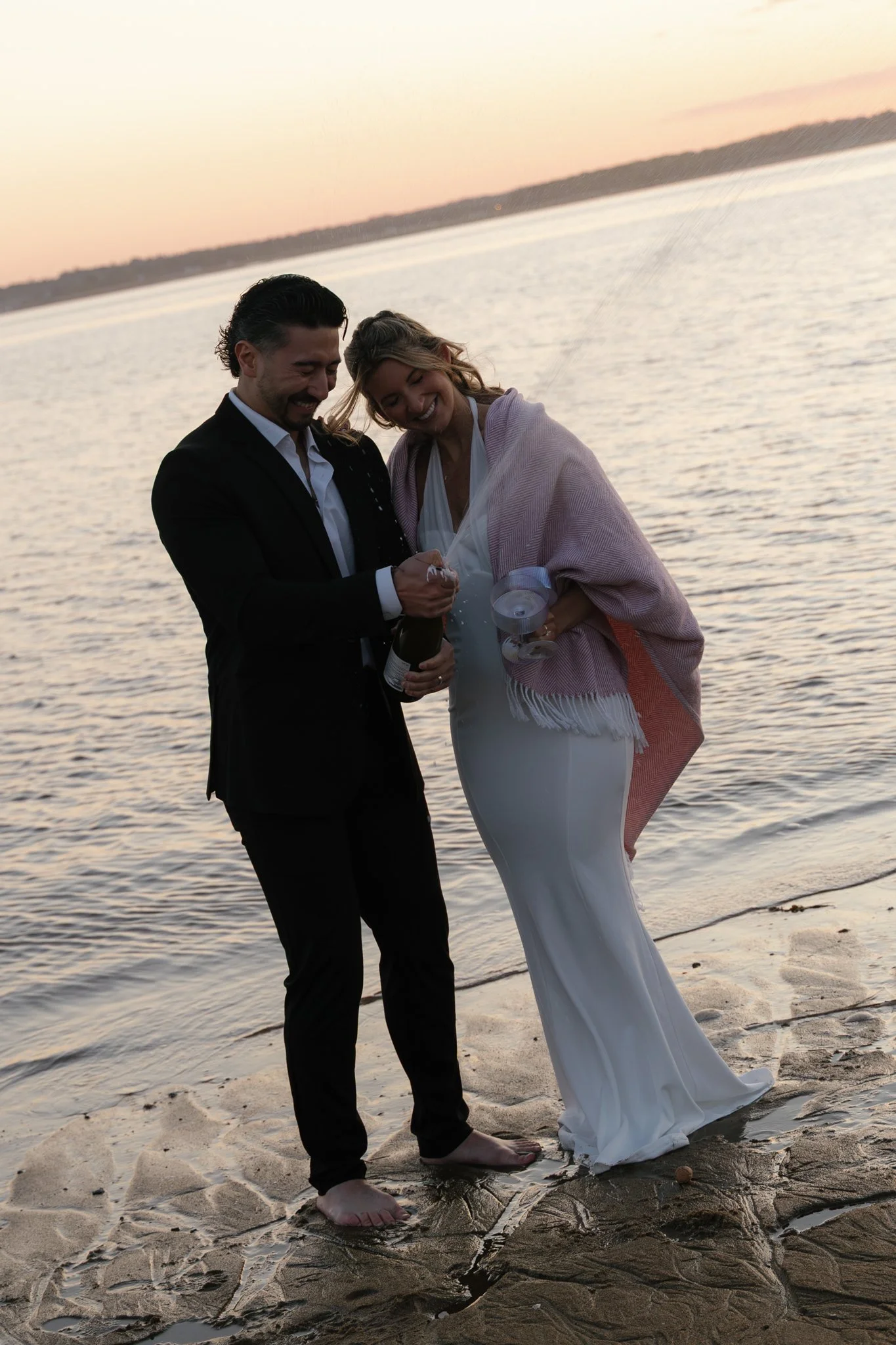 A couple dressed in formal attire, possibly a bride and groom, standing barefoot on the beach at sunset, celebrating with champagne.