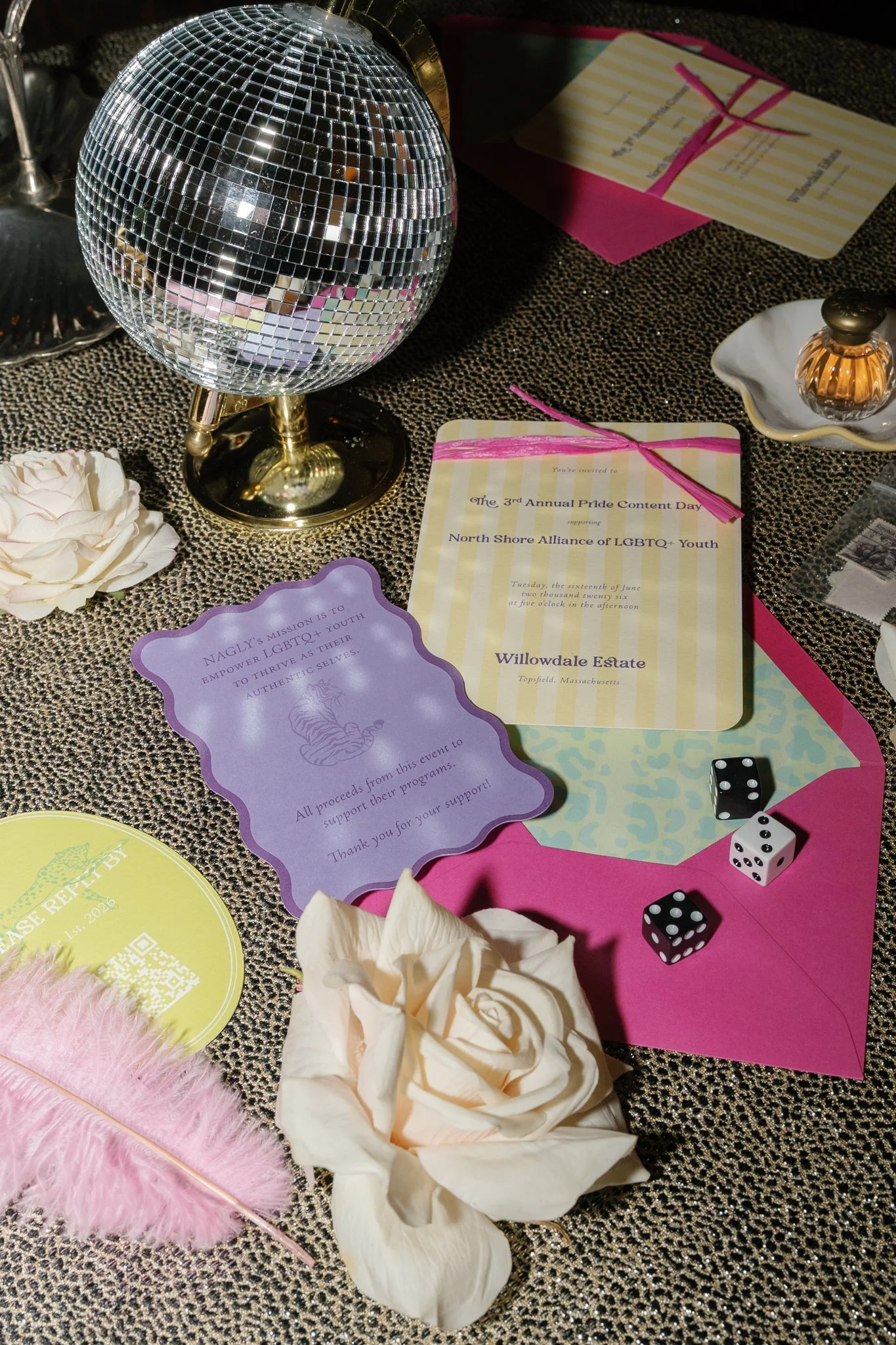 Disco ball, pink feather, white rose, small decorative bottle, game dice, and event invitations on a textured tablecloth at a Pride celebration or event.