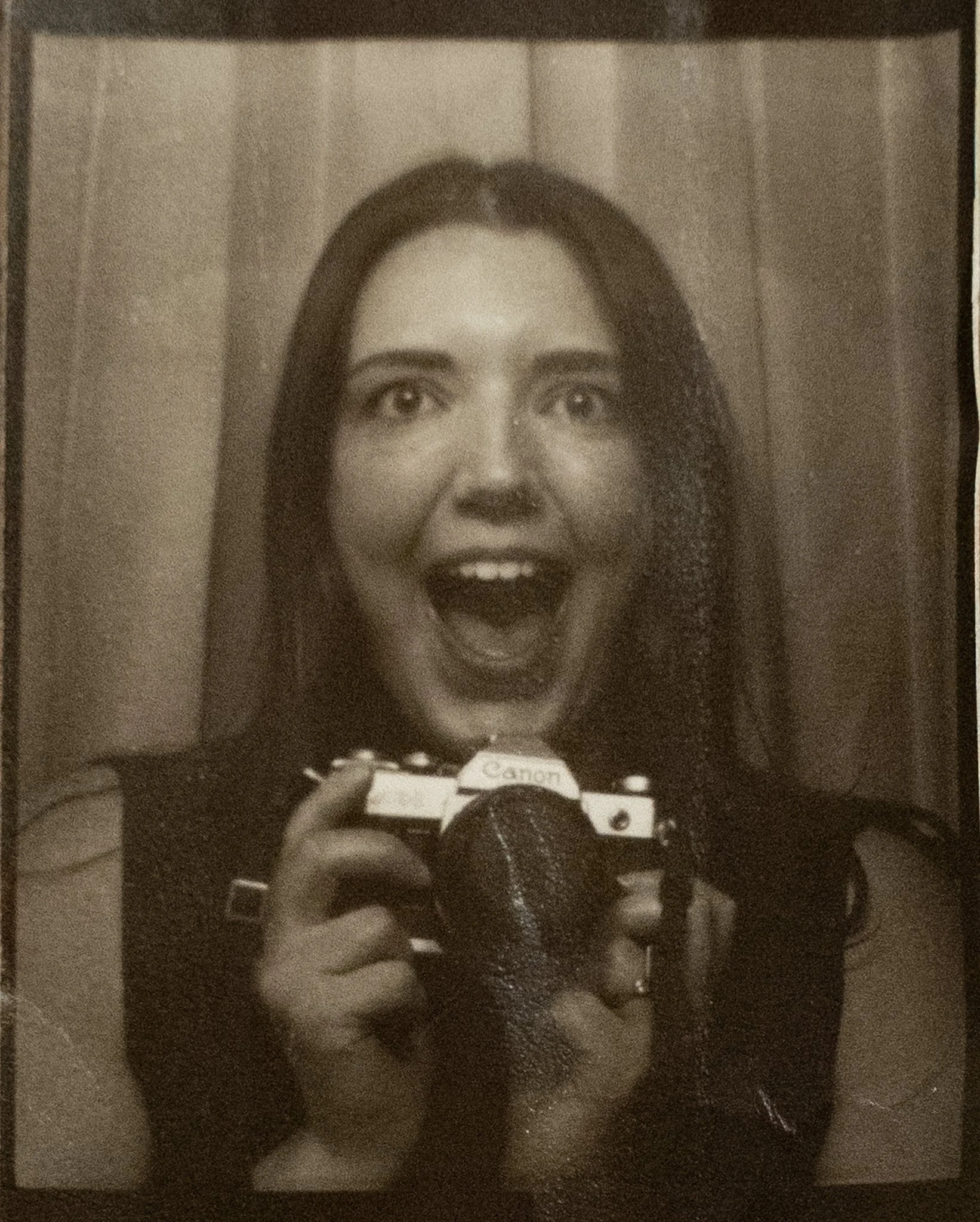A woman with long dark hair taking a mirror selfie with a camera, smiling with wide eyes and open mouth.