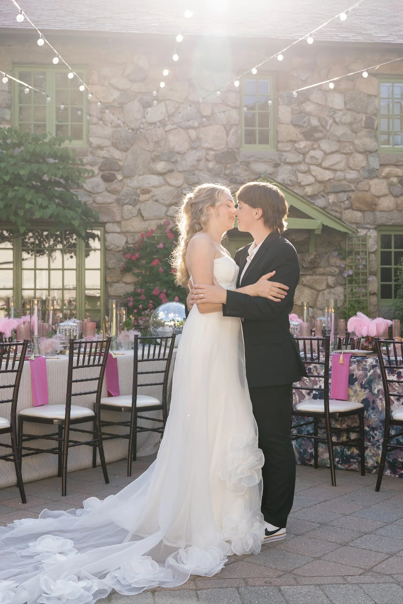 A bride in a white wedding dress and groom in a black suit sharing a dance outdoors at a wedding with decorated tables and string lights.