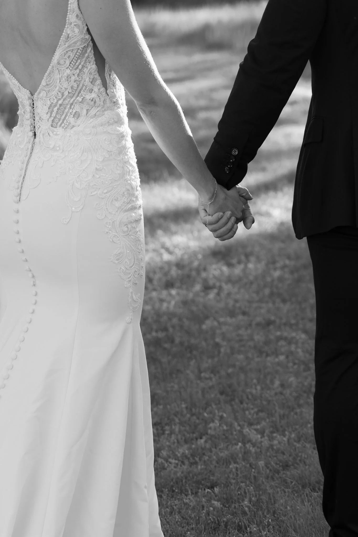Close-up of a bride and groom holding hands, the bride wears a white lace wedding gown with a zipper and buttons at the back, the groom wears a black suit, outdoors on a grassy field.