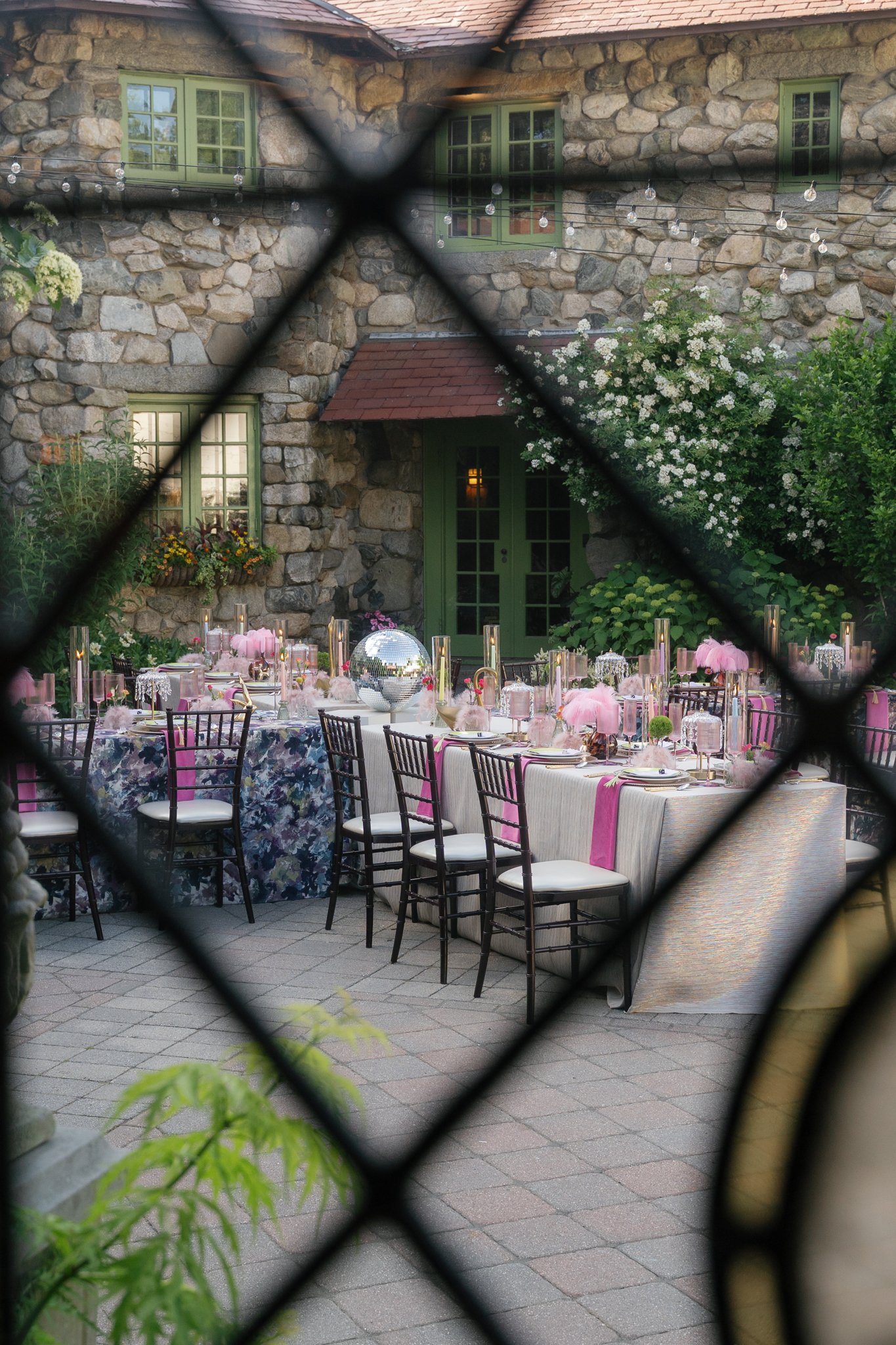 A outdoor party setup with tables, chairs, and decorations, seen through a black diamond-shaped fence with green foliage in the foreground. The background features a stone house with green window frames and string lights hanging between the windows.