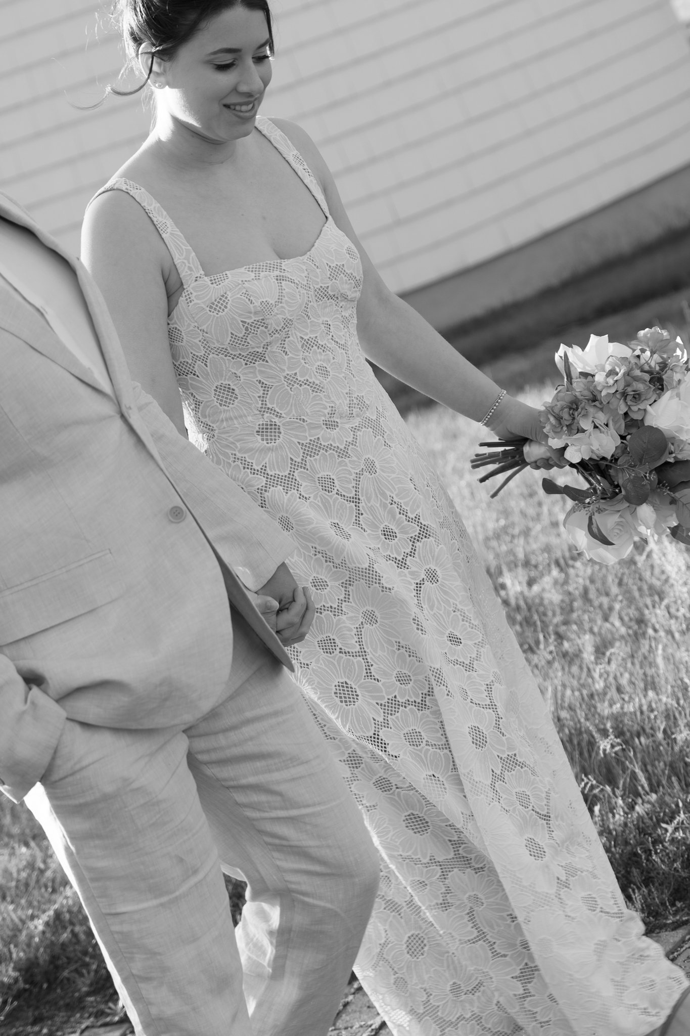 A woman in a lace dress holding a bouquet of flowers, standing outdoors next to a man in a light-colored suit.