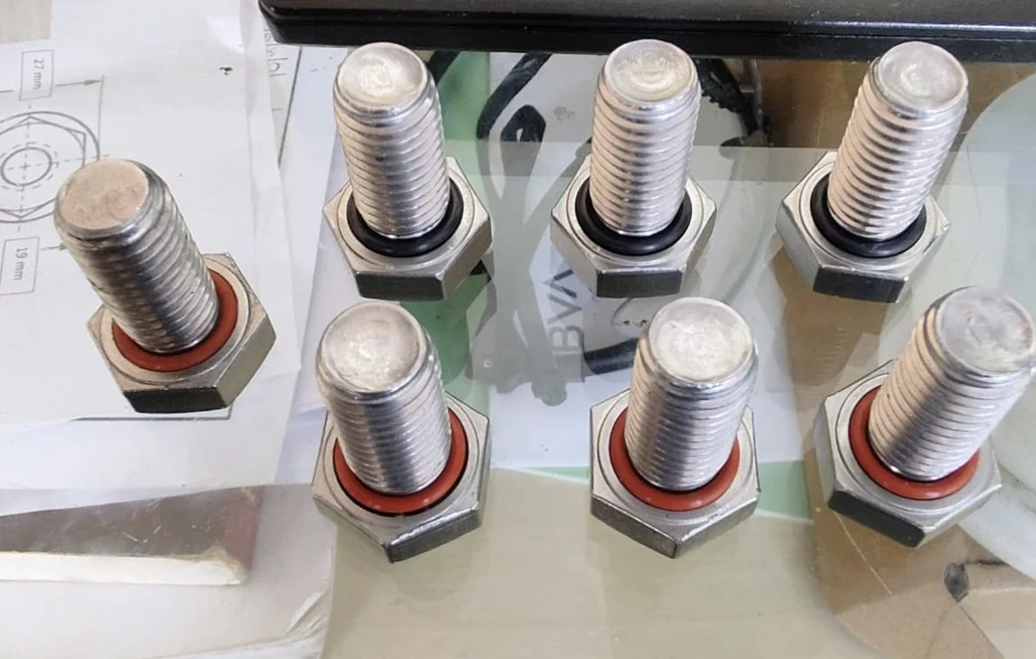 Siete tornillos grandes sobre una superficie de vidrio, algunos con juntas de goma rojas.