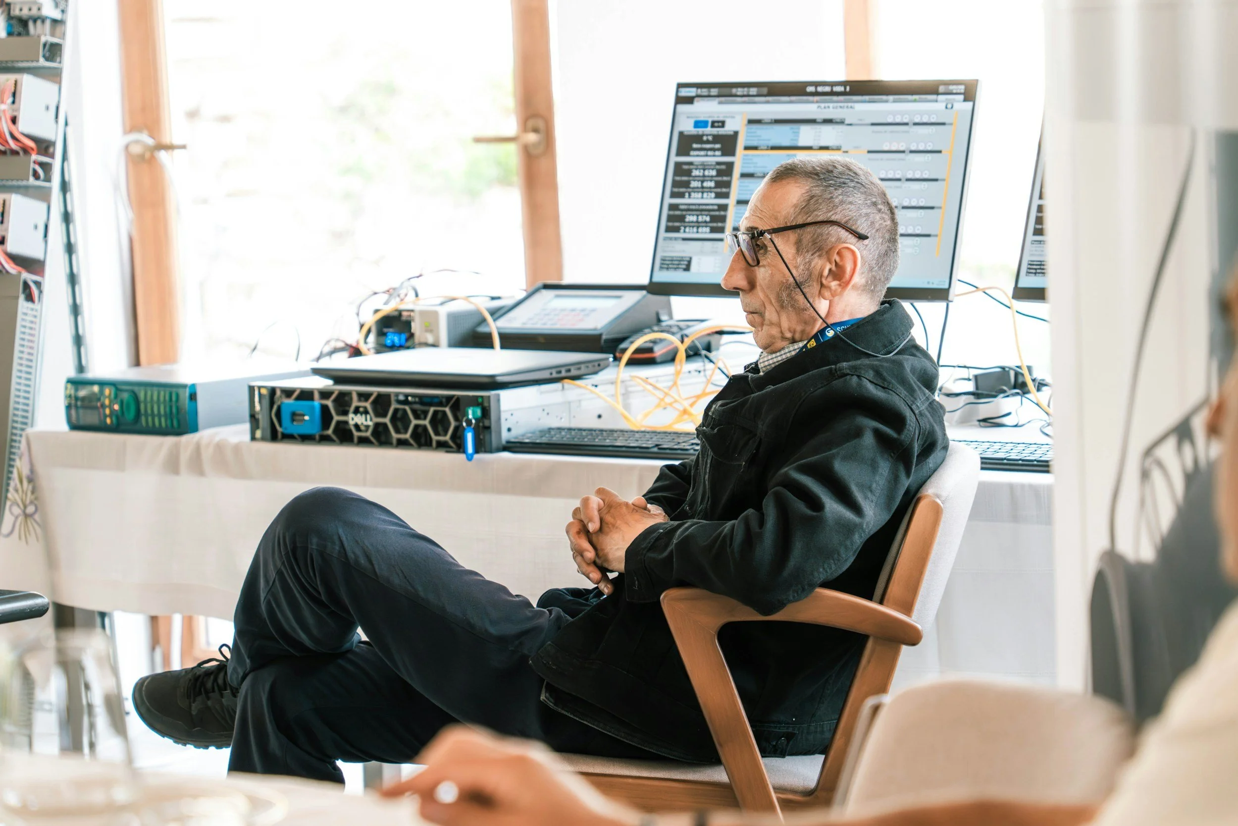 Hombre sentado en una silla, con gafas, en un entorno de oficina, mirando una pantalla de computadora grande que muestra gráficos y datos, rodeado de cables y equipo tecnológico.