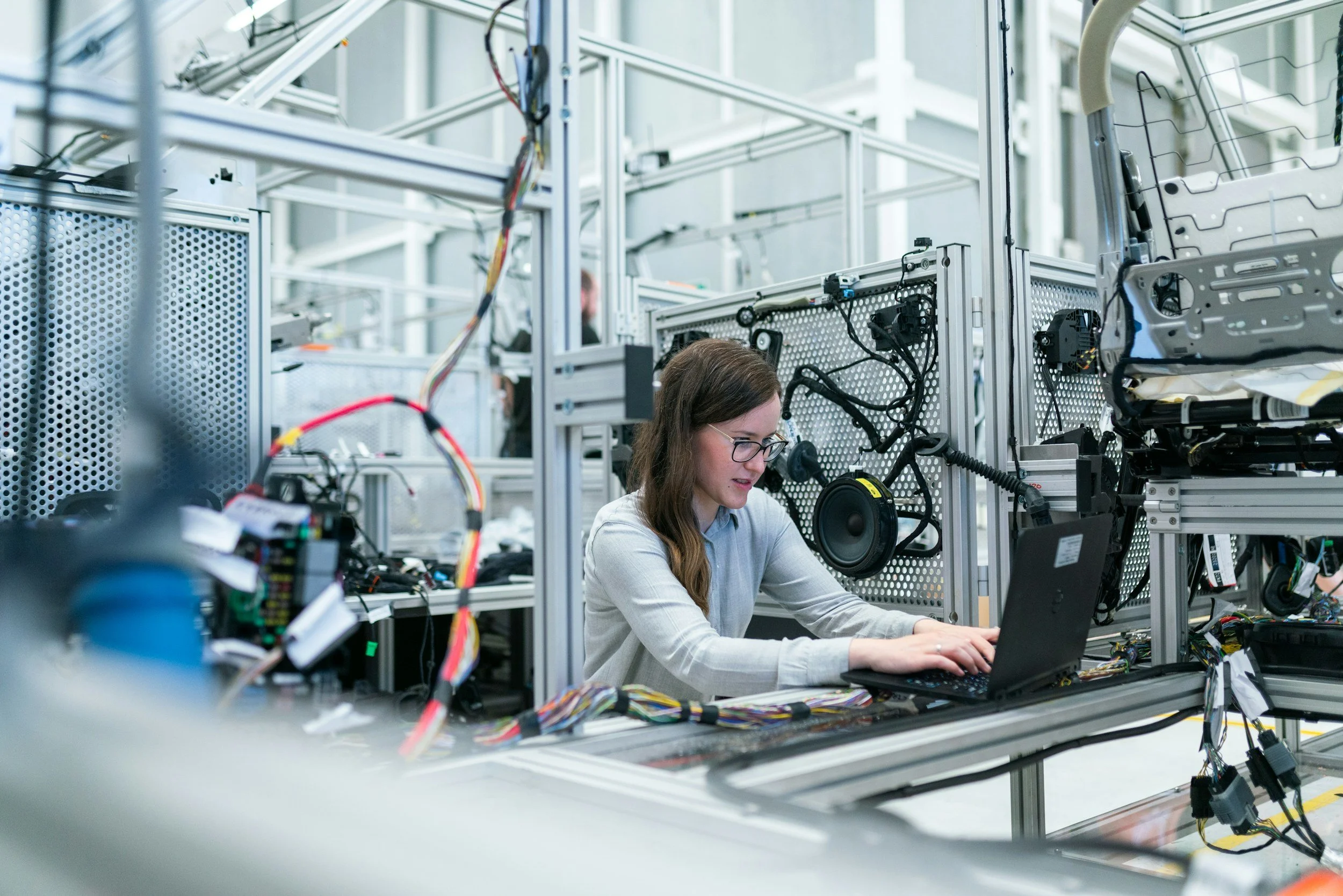Mujer trabajando en un laboratorio de ingeniería con un ordenador portátil y componentes electrónicos en un entorno con estructura de metal y cables.