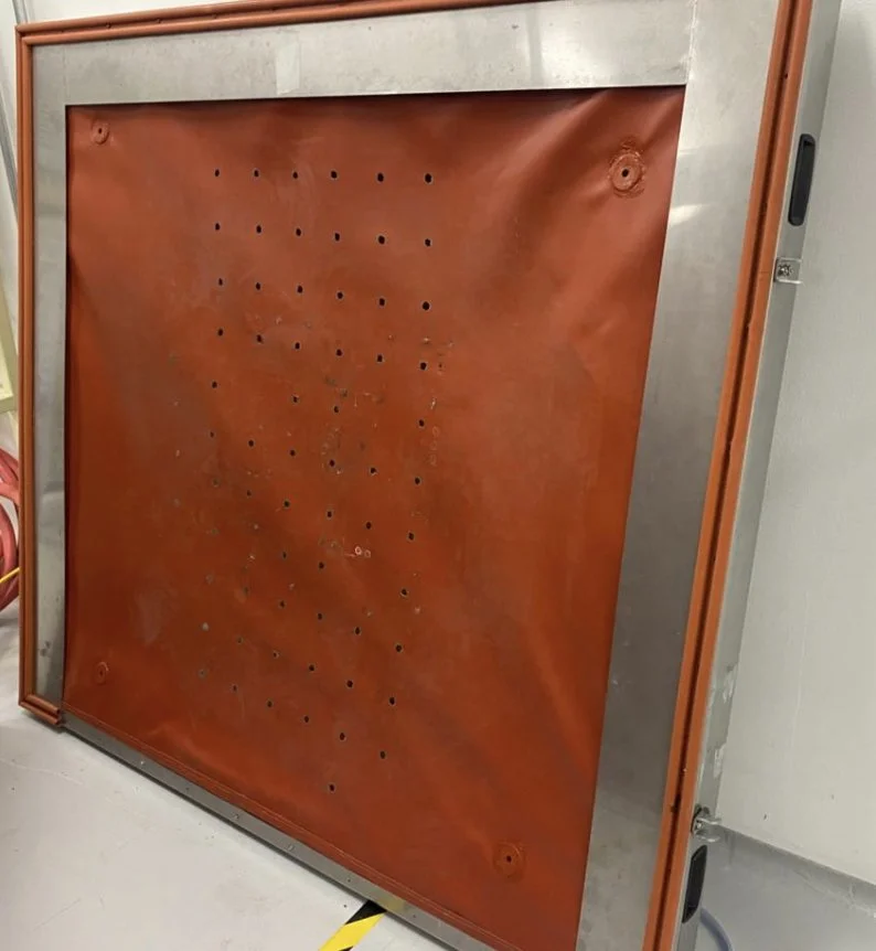 Una pizarra de color naranja con agujeros distribuidos en un patrón, colocada sobre un soporte metálico y enchufes eléctricos visibles a la izquierda.