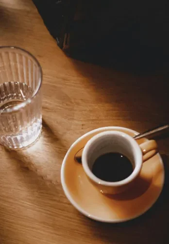 Cup of black coffee on a saucer, a glass of water, and a black bag on a wooden table.