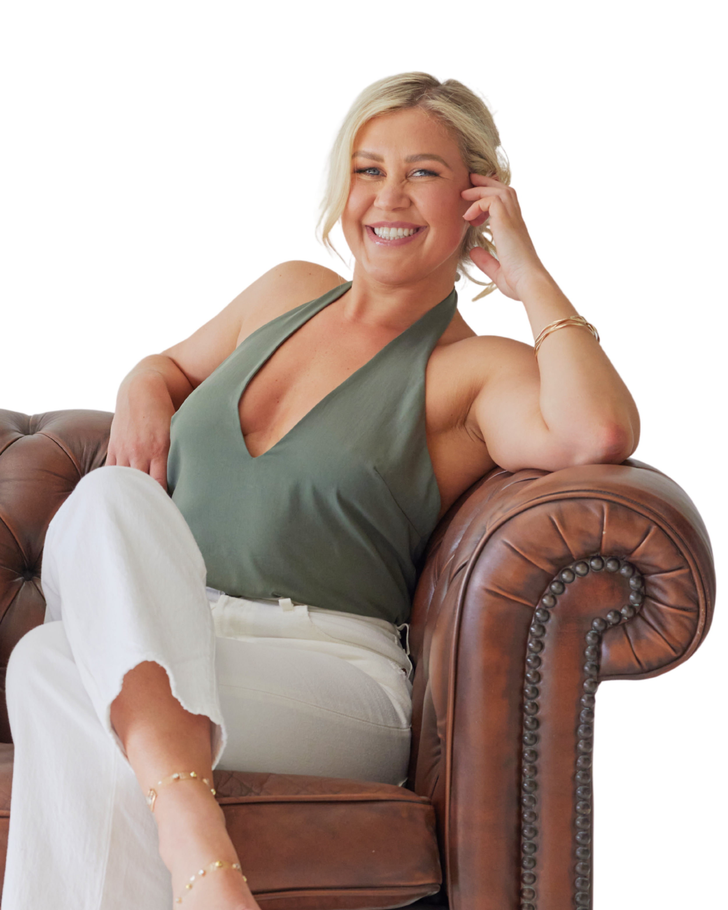 A woman with blonde hair sitting on a brown leather armchair, smiling and looking at the camera. She is wearing a dark green sleeveless top, white pants, and jewellery on her arm. She is warm, confident, a teacher and someone trustworthy.