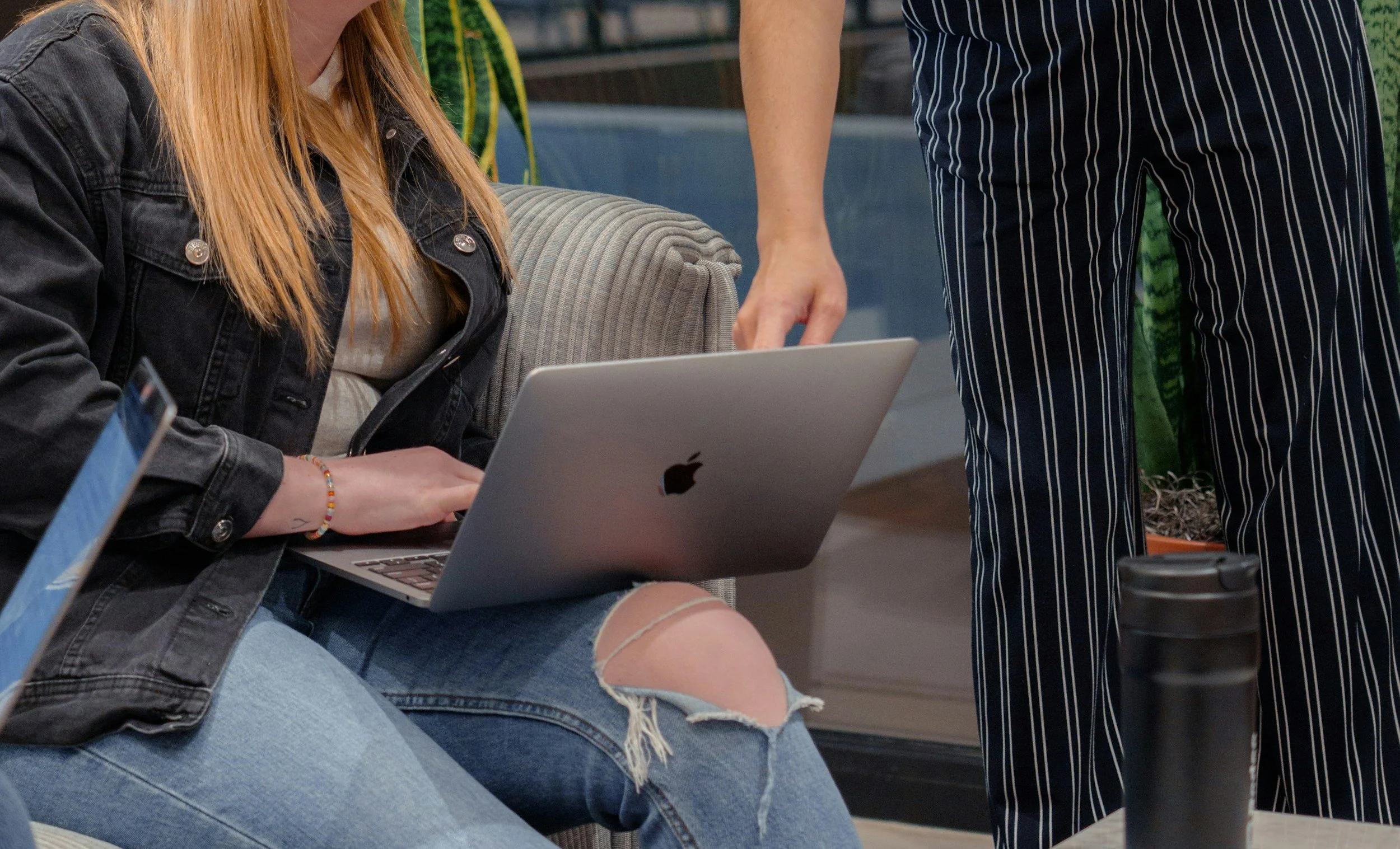 A woman with long red hair wearing a black denim jacket and ripped jeans sitting at a table with a silver MacBook laptop. Two other people are standing nearby, one in striped pants and another with a green patterned shirt, in a setting with plants and a modern interior.